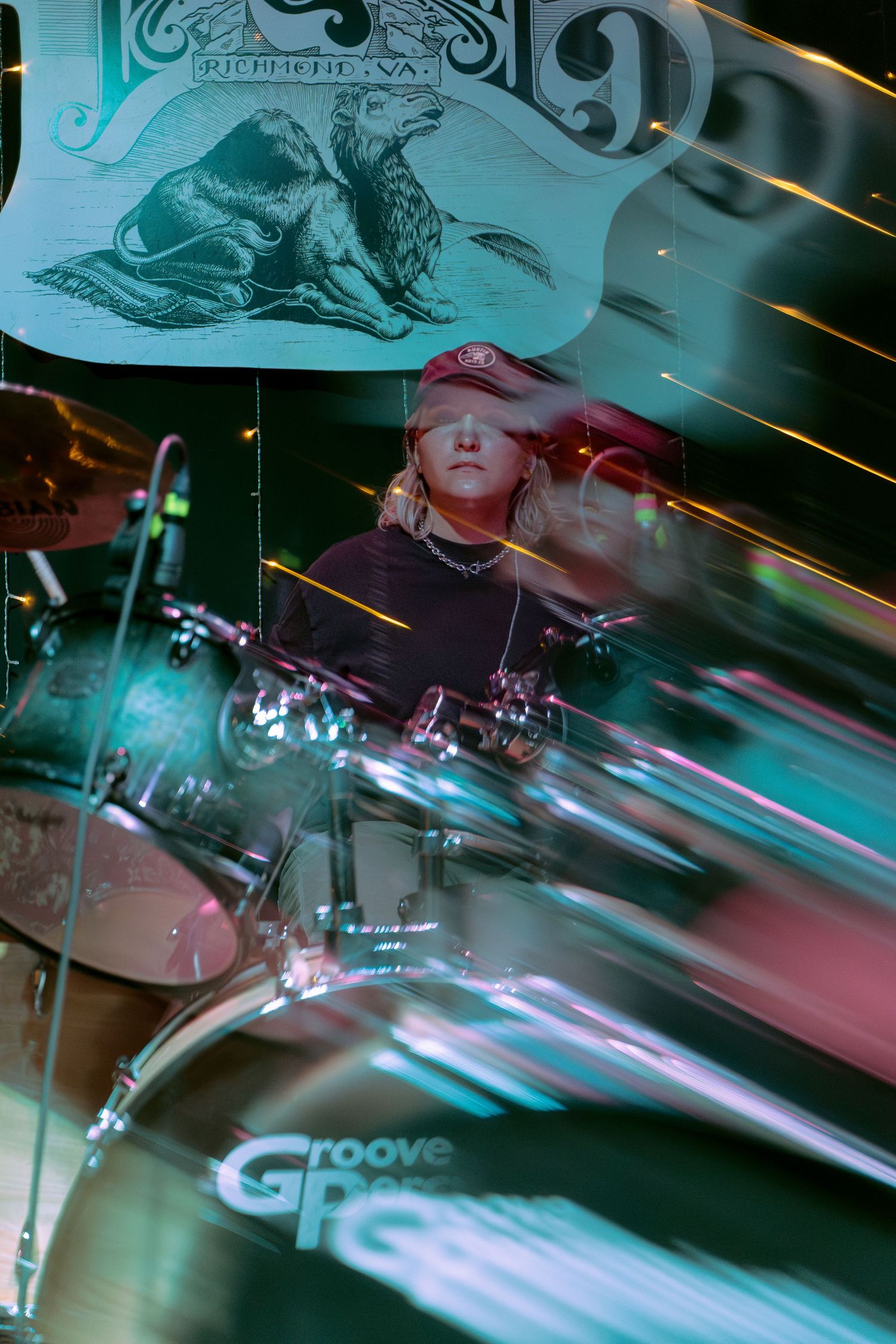Motion blur photo of cymbals and drum equipment shown in turquoise lighting at a live music performance.