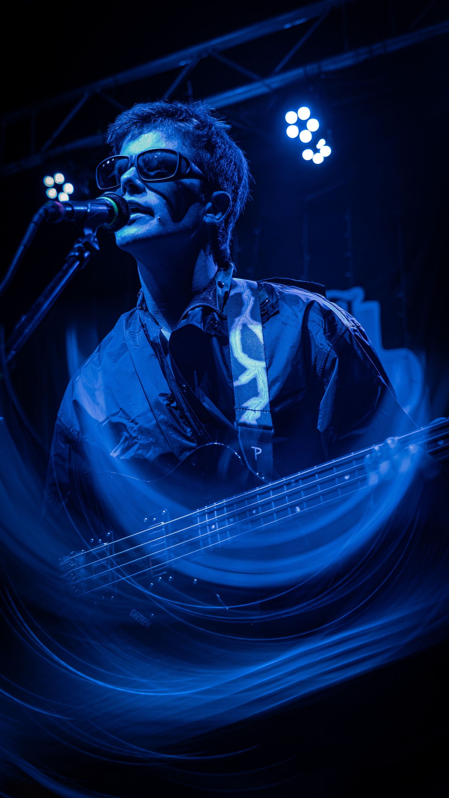 A performer in blue lighting sings into a microphone while playing bass guitar on stage during a concert performance.