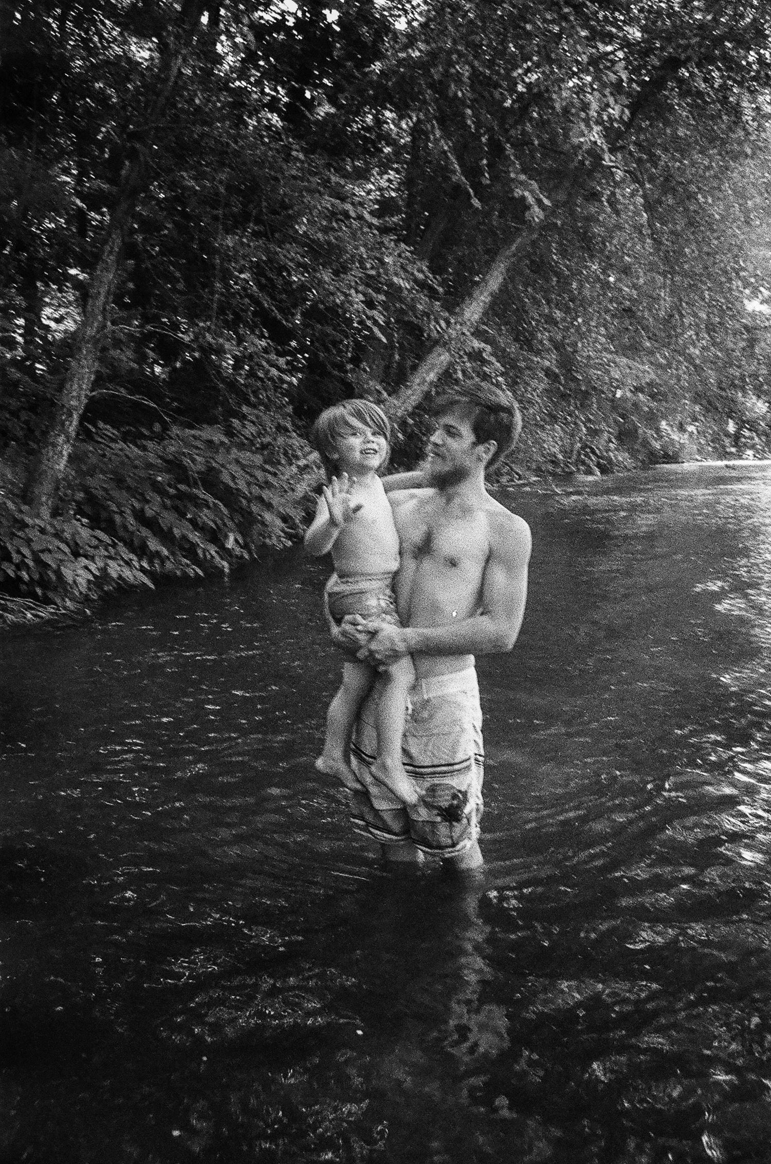 A parent holding a small child while standing in a flowing stream on a summer day.