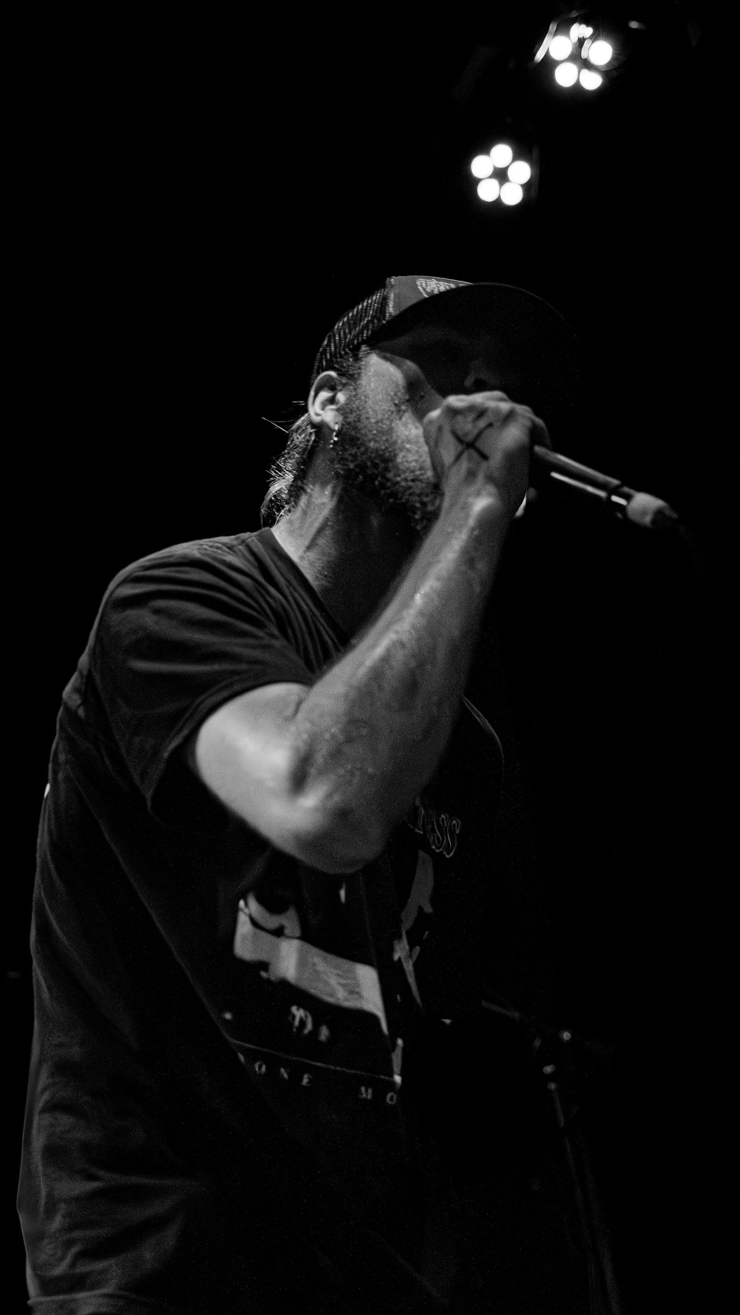 Silhouetted performer in black holds a microphone during a dark concert performance on stage with white lights in background.