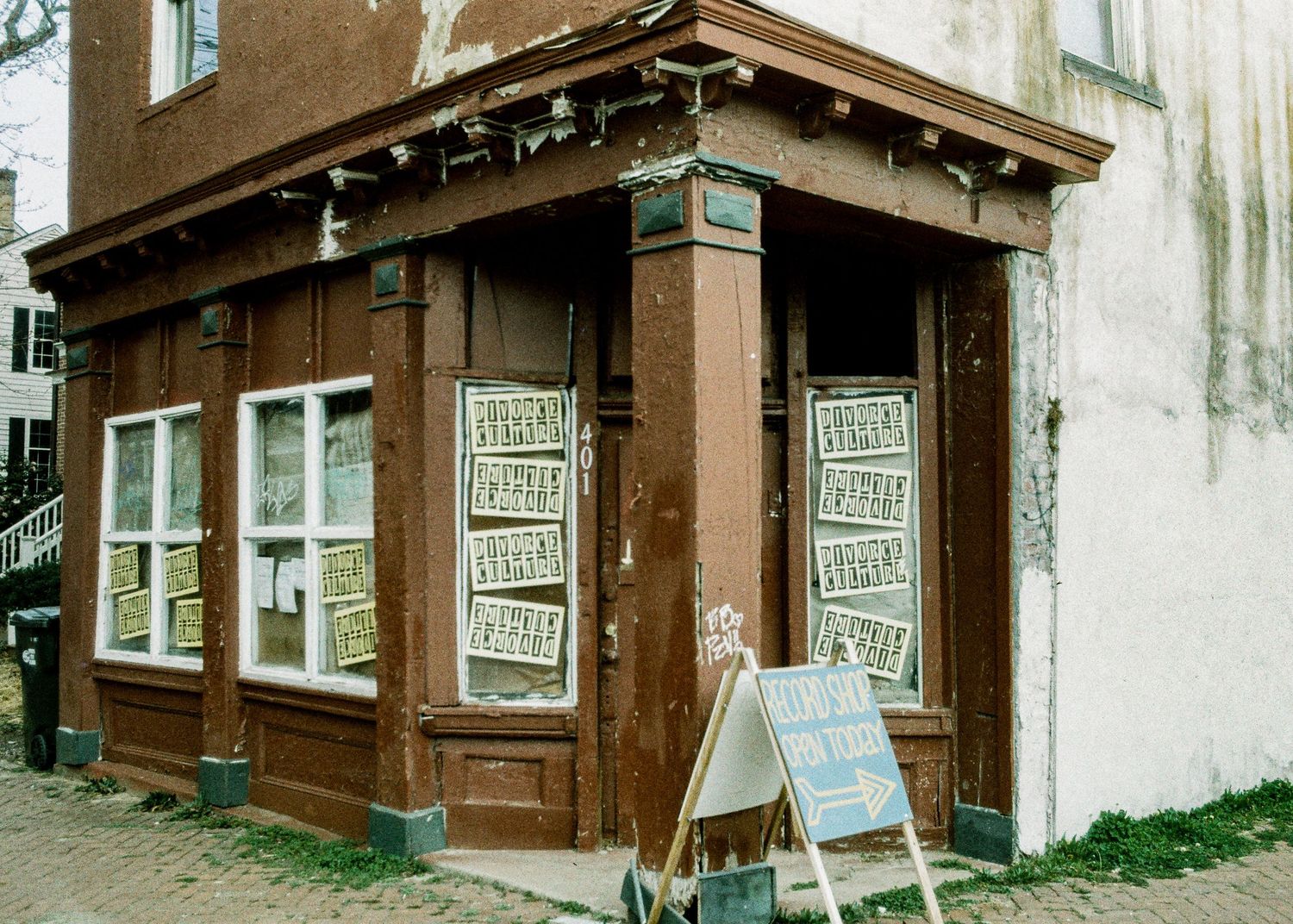 Historic brown wooden storefront with multiple windows and signs sits against an aged white wall on a cobblestone street.