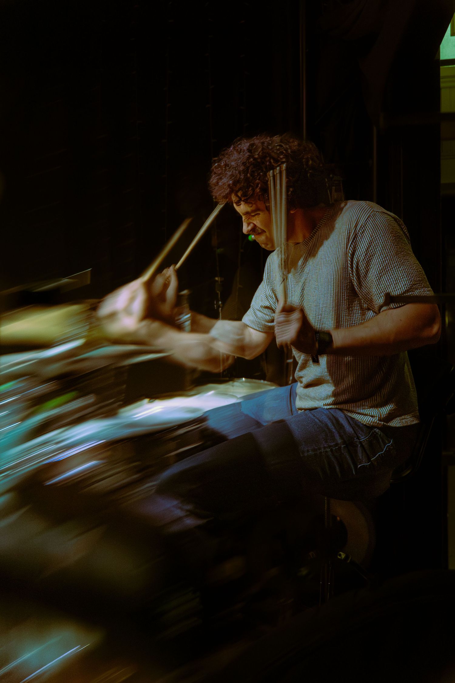 Drummer with curly hair performs a dynamic drum solo in low light conditions, captured in motion blur sequence.