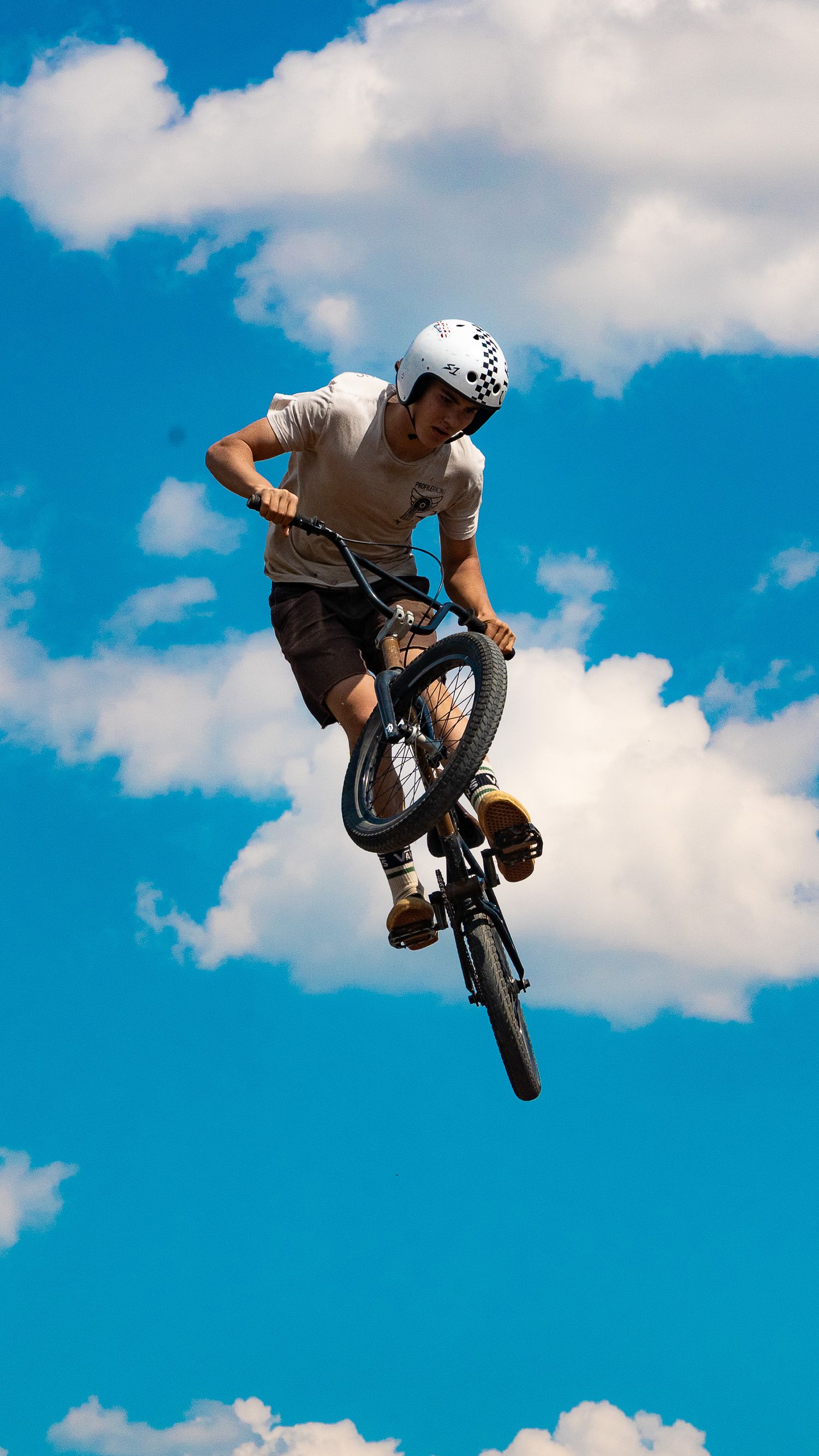 Athlete performs aerial BMX trick with bike against bright blue sky and puffy white clouds.