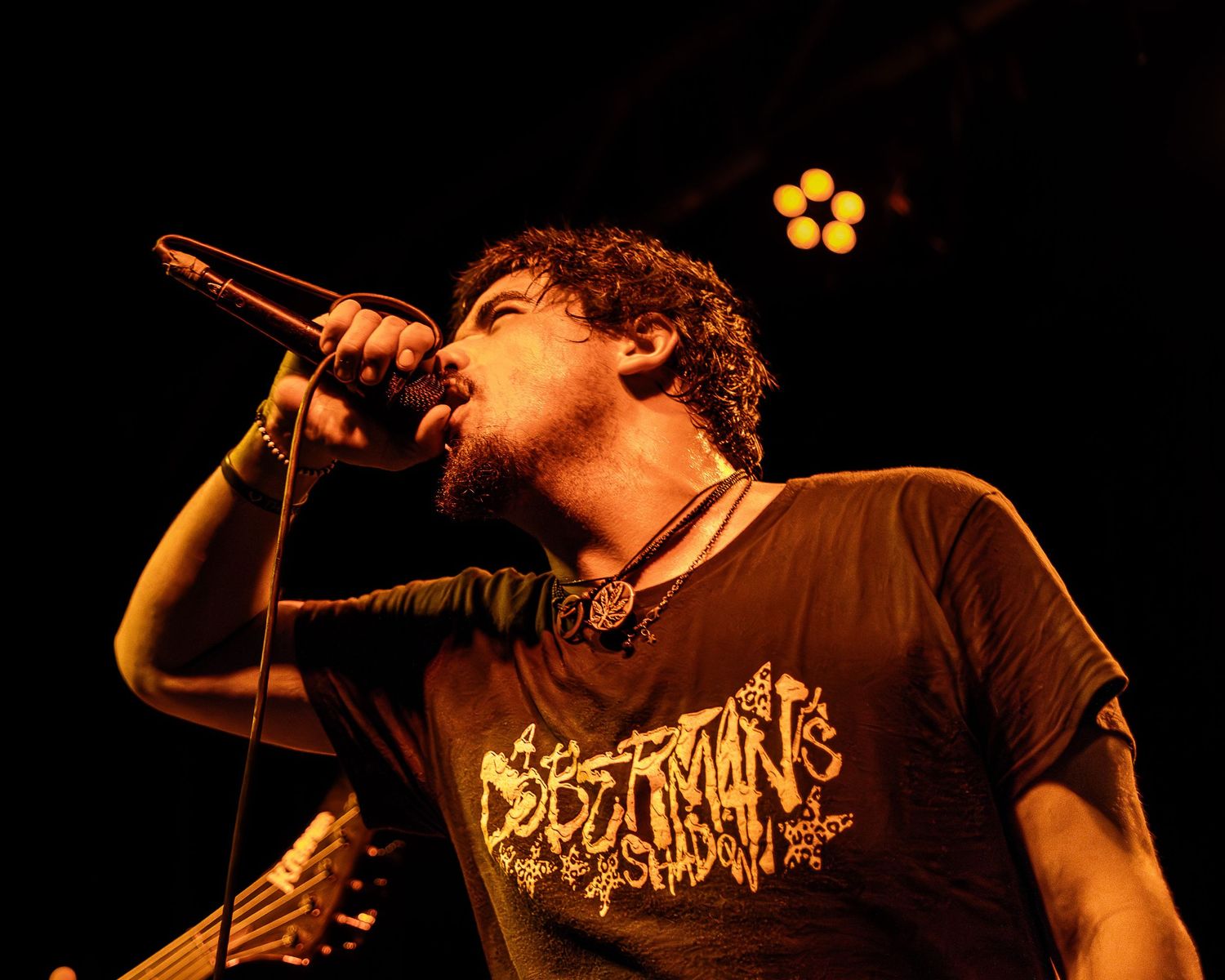 A performer in a brown t-shirt sings passionately into a microphone on stage with warm lighting in the background.