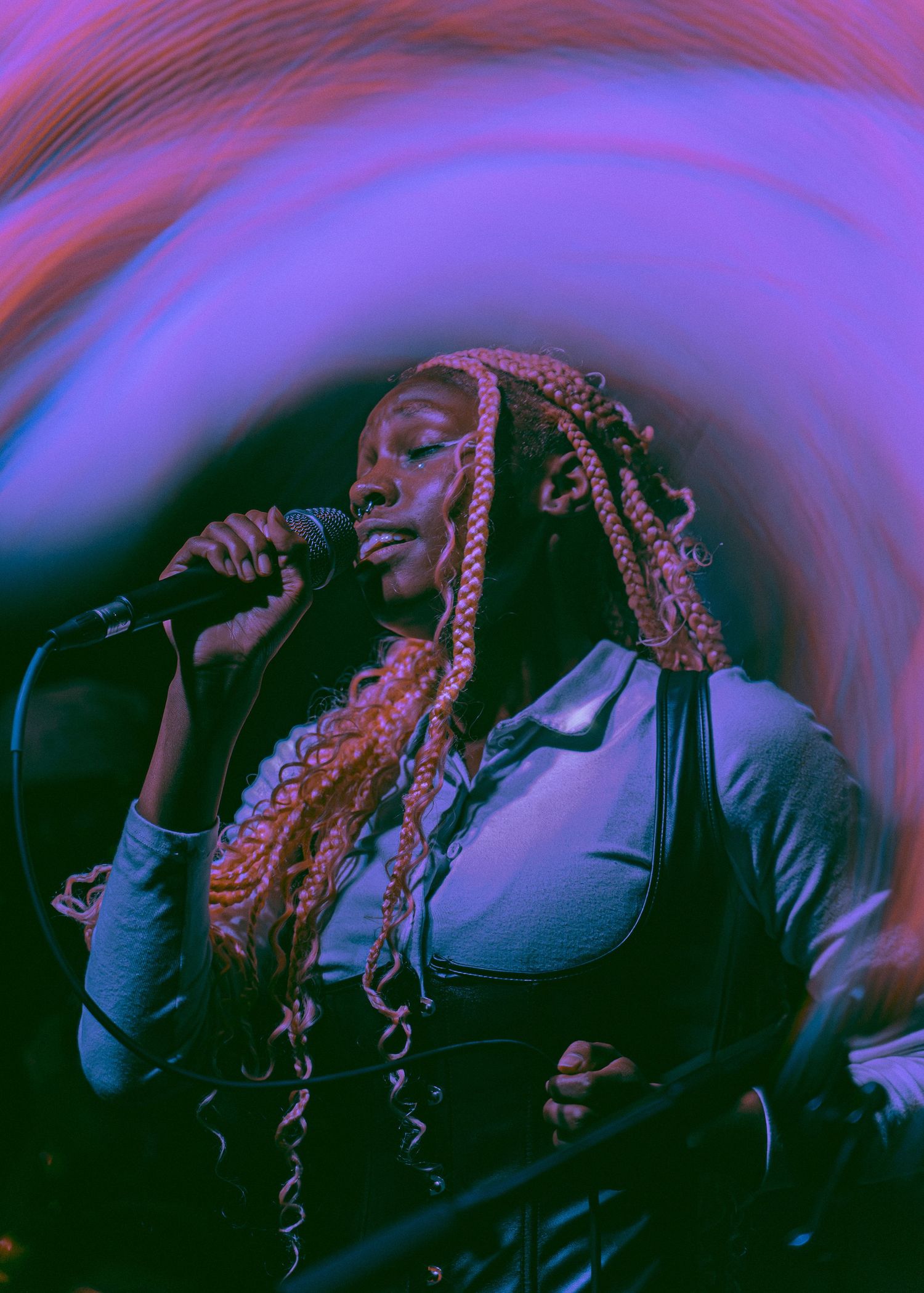 A performer with long braids singing into a microphone against a purple and blue swirled background.
