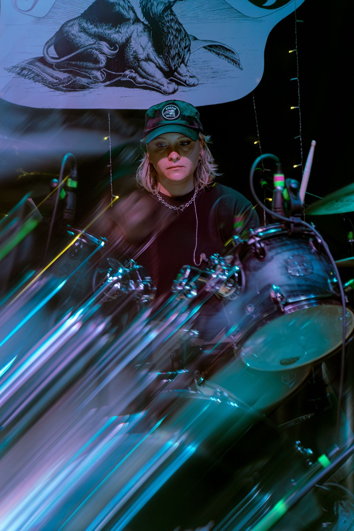 Concert drummer performs behind drum kit under blue and green stage lighting with motion blur effects.