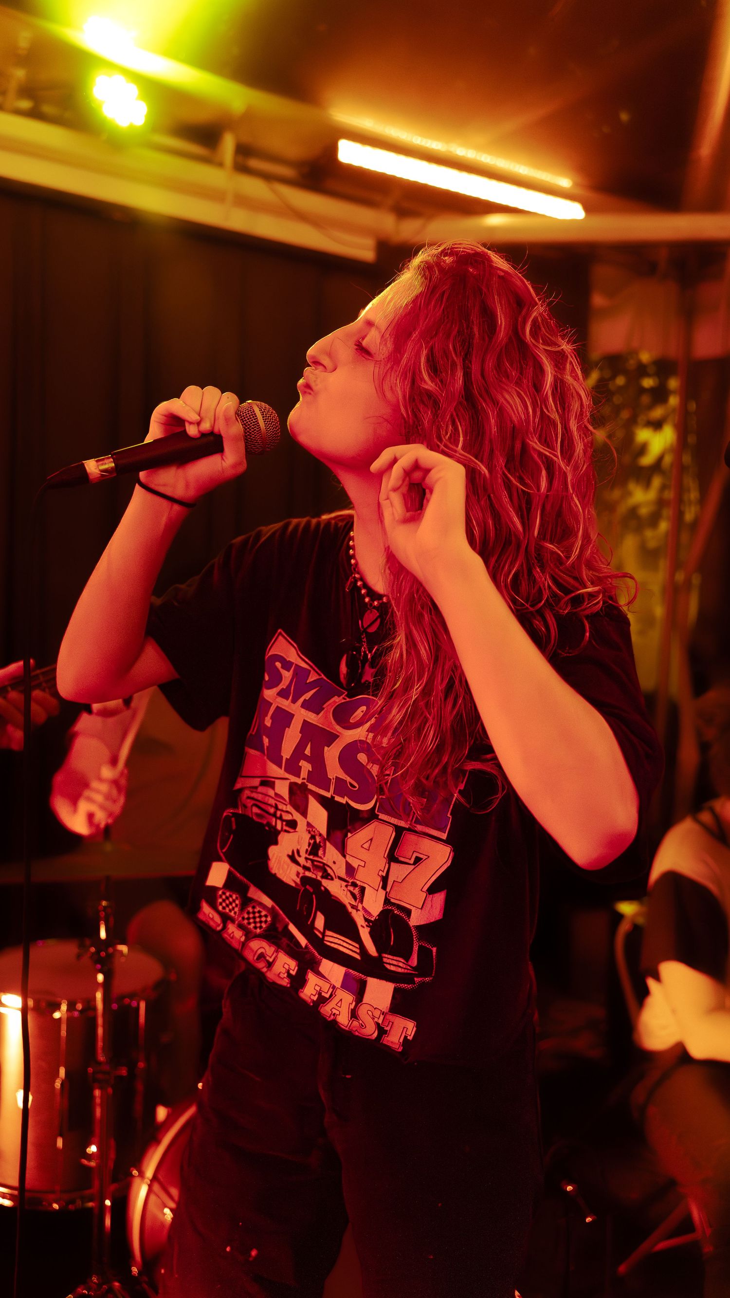 A metal singer with bright red hair performs passionately in a dimly lit venue with dramatic red stage lighting.