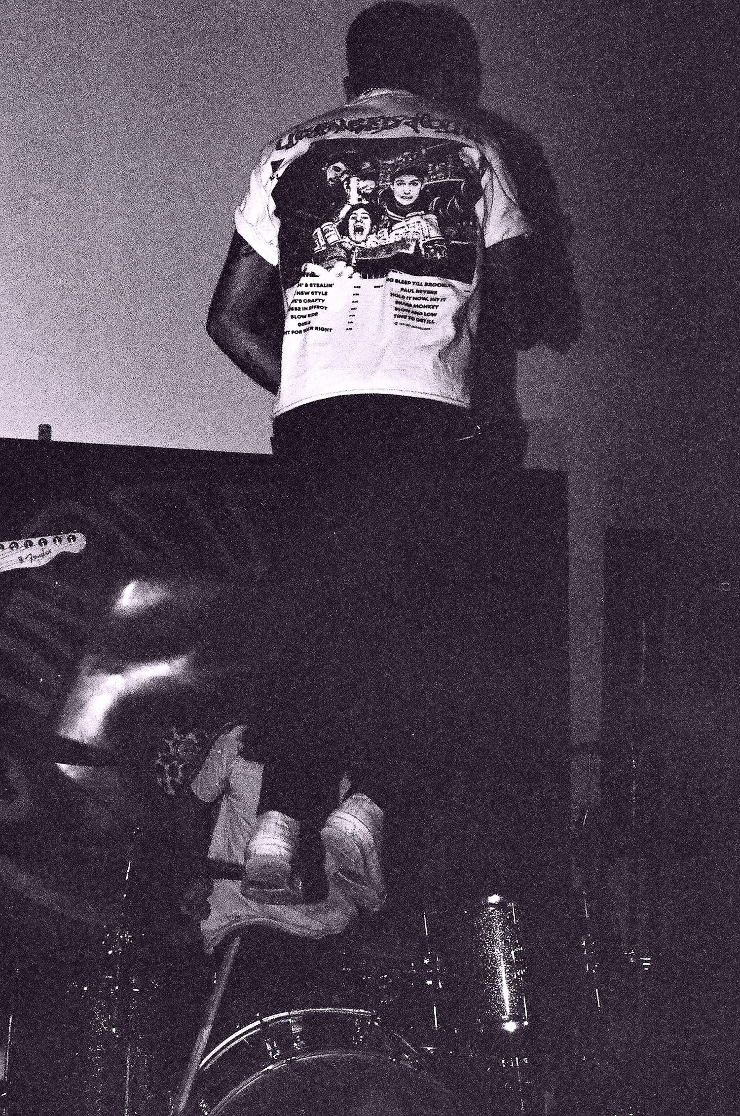 Black and white concert photo showing performer wearing band t-shirt from behind on stage.