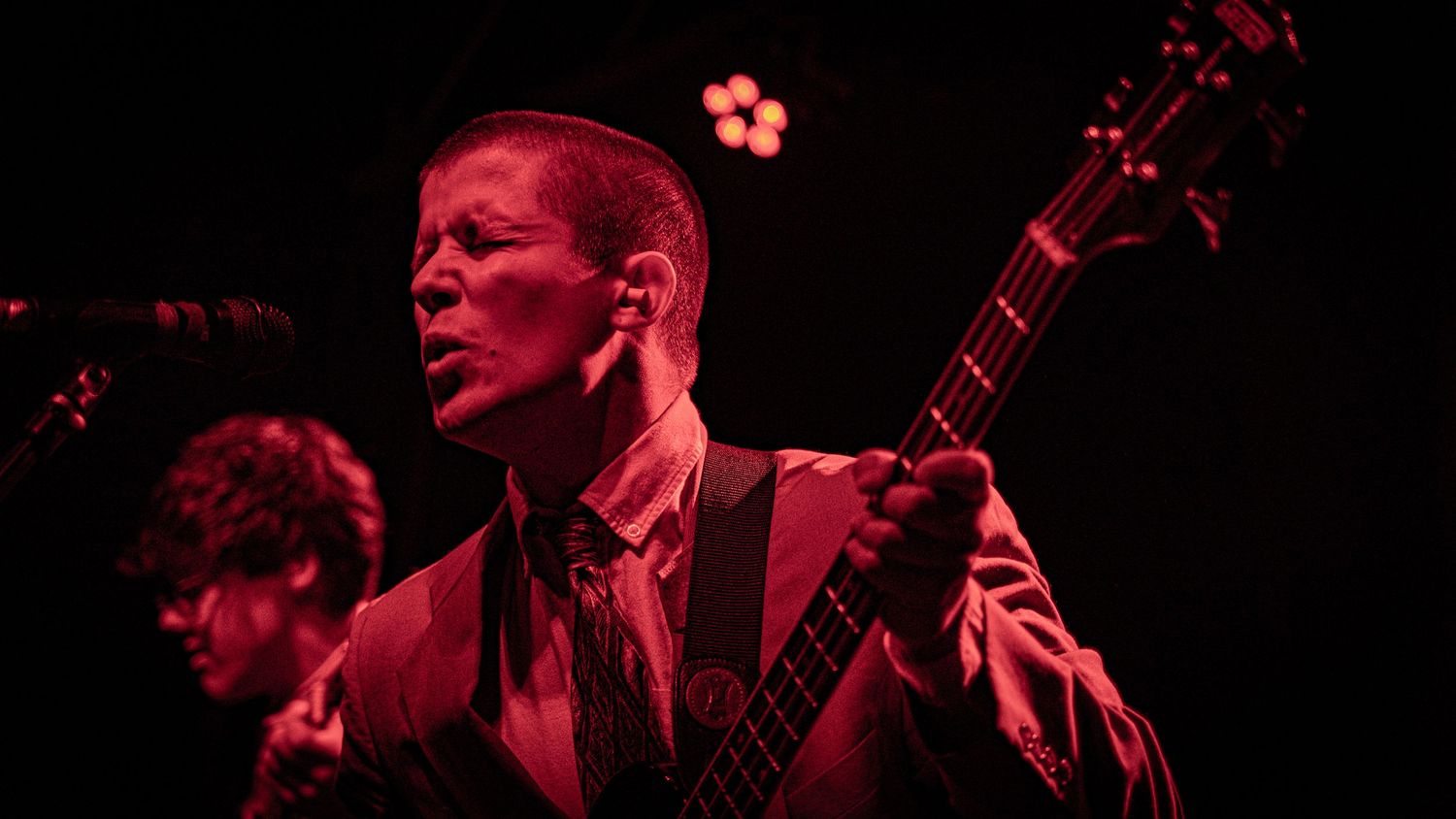 Musician playing guitar on darkened stage illuminated by dramatic red spotlights.
