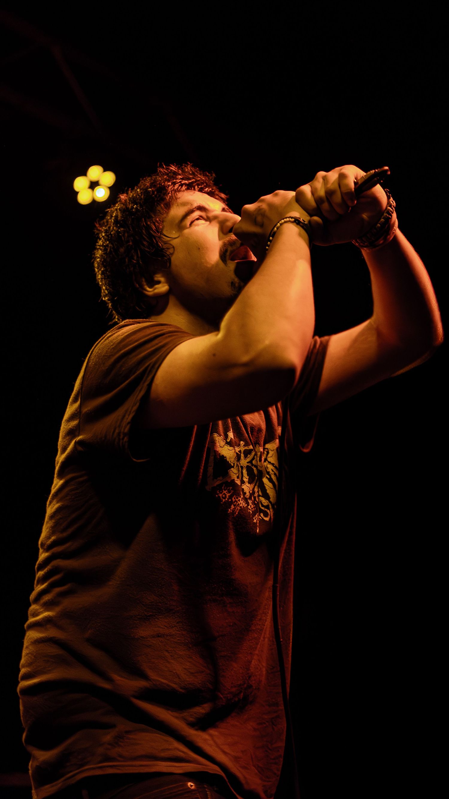 A performer sings passionately into a microphone on a dark stage with dramatic backlighting silhouetting their figure.