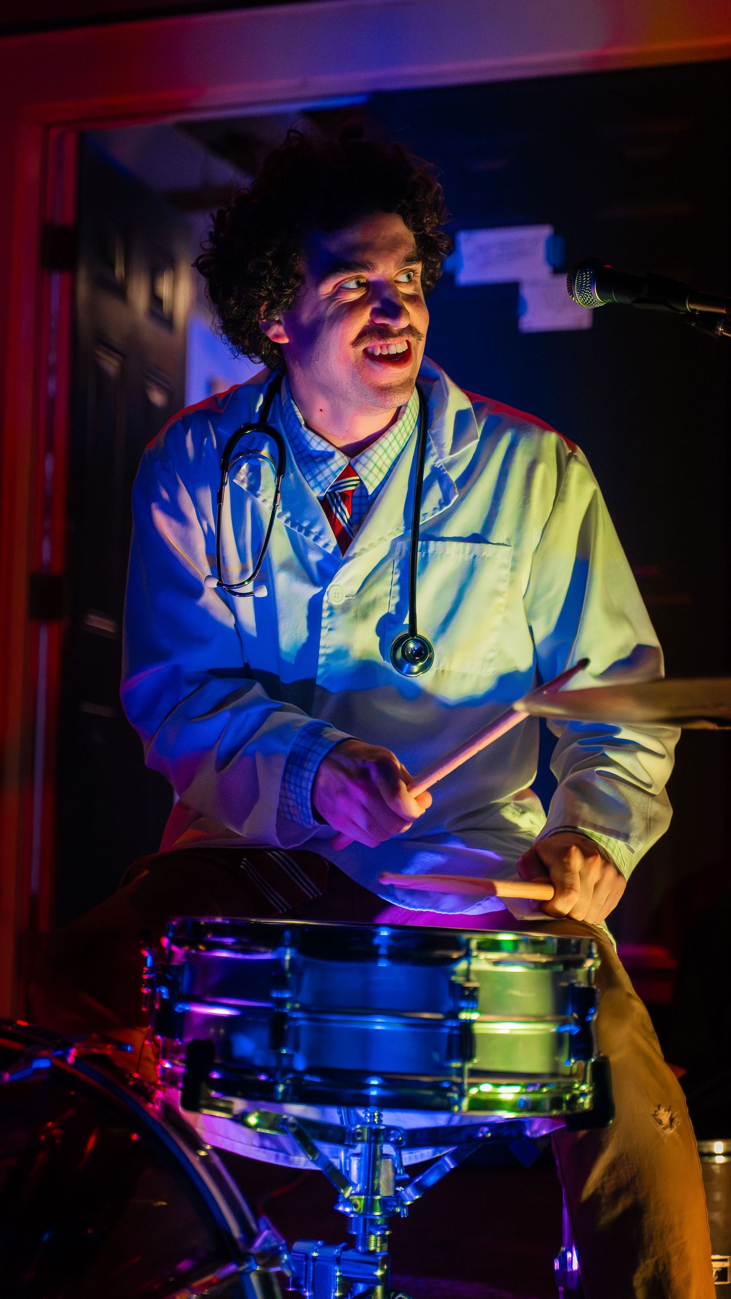 Drummer playing at a drum set with colorful stage lighting creating dramatic shadows.