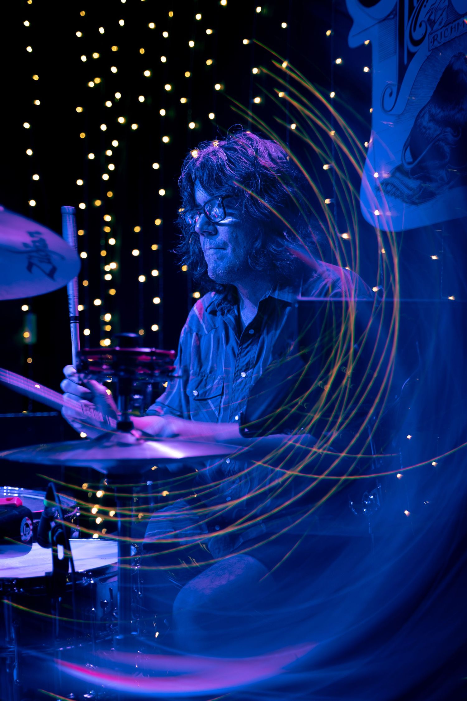 Drummer performs in dark blue lighting with swirling light trails and sparkles creating a mesmerizing nighttime atmosphere.