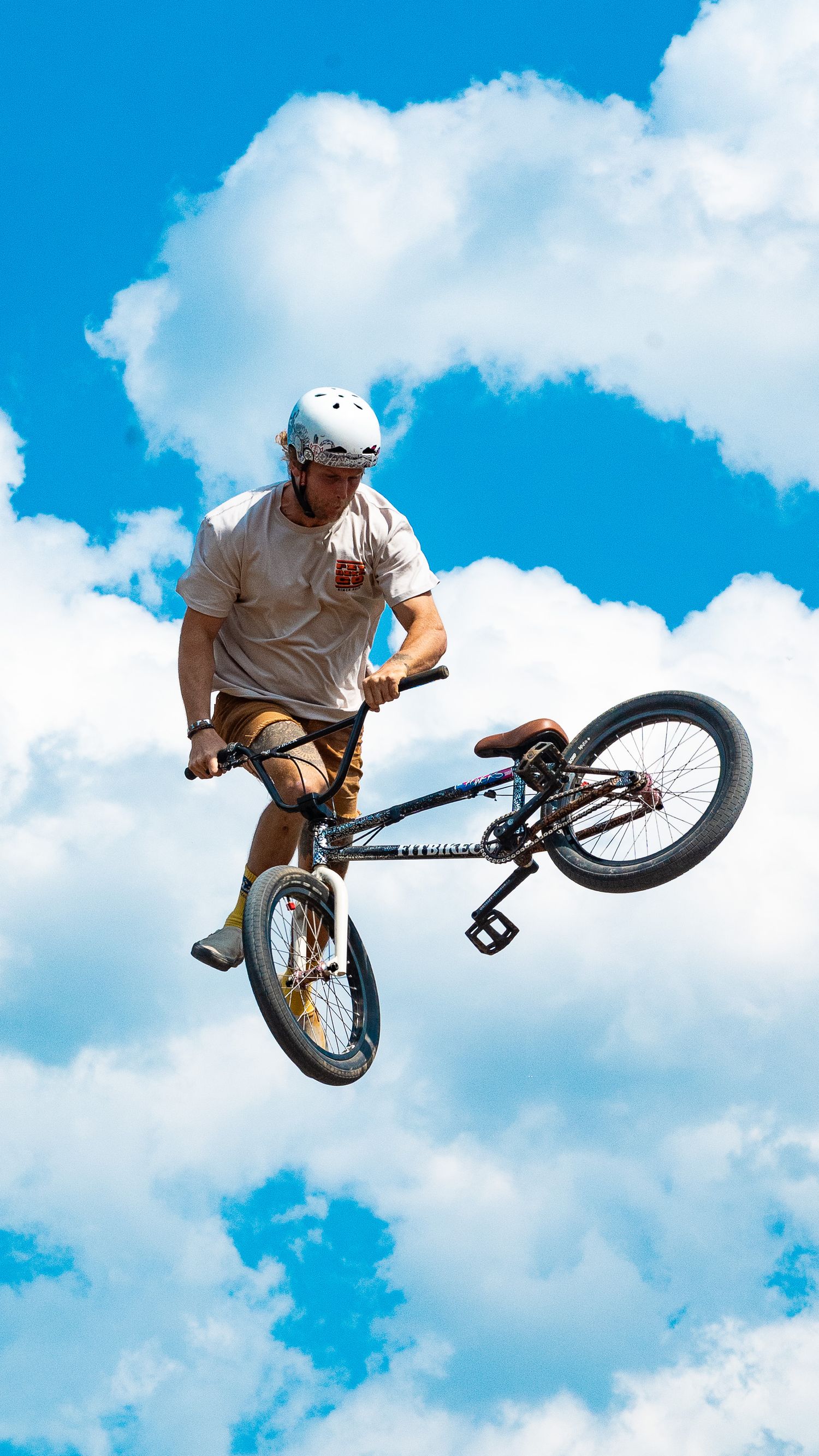 Airborne BMX rider performs stunt with bike tilted sideways against blue sky with fluffy clouds.
