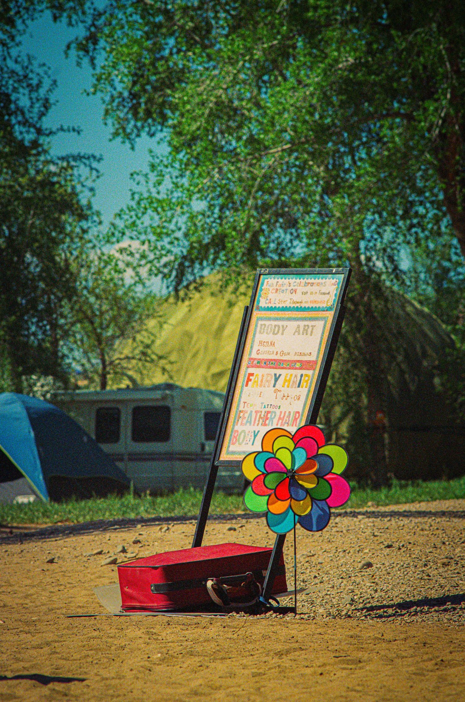 Colorful pinwheel flower and sign board next to a red case on dirt ground with trees in background.