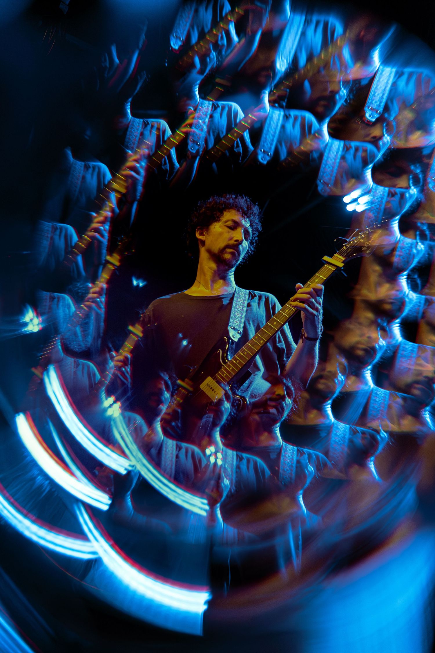A guitarist performs on stage with swirling blue light trails creating a dynamic long exposure effect in the background.