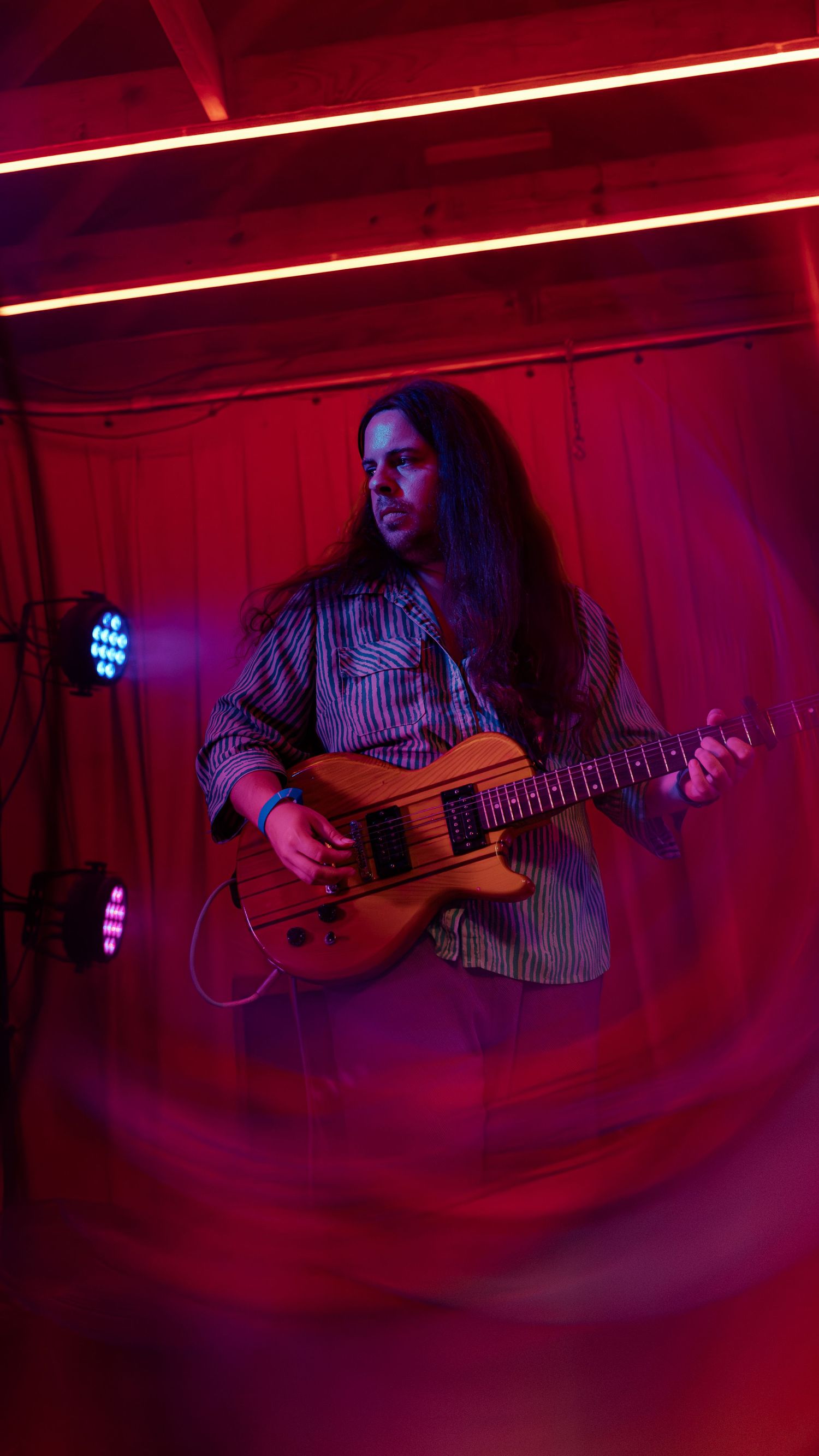 A musician plays acoustic guitar under dramatic red stage lighting creating a moody performance atmosphere.