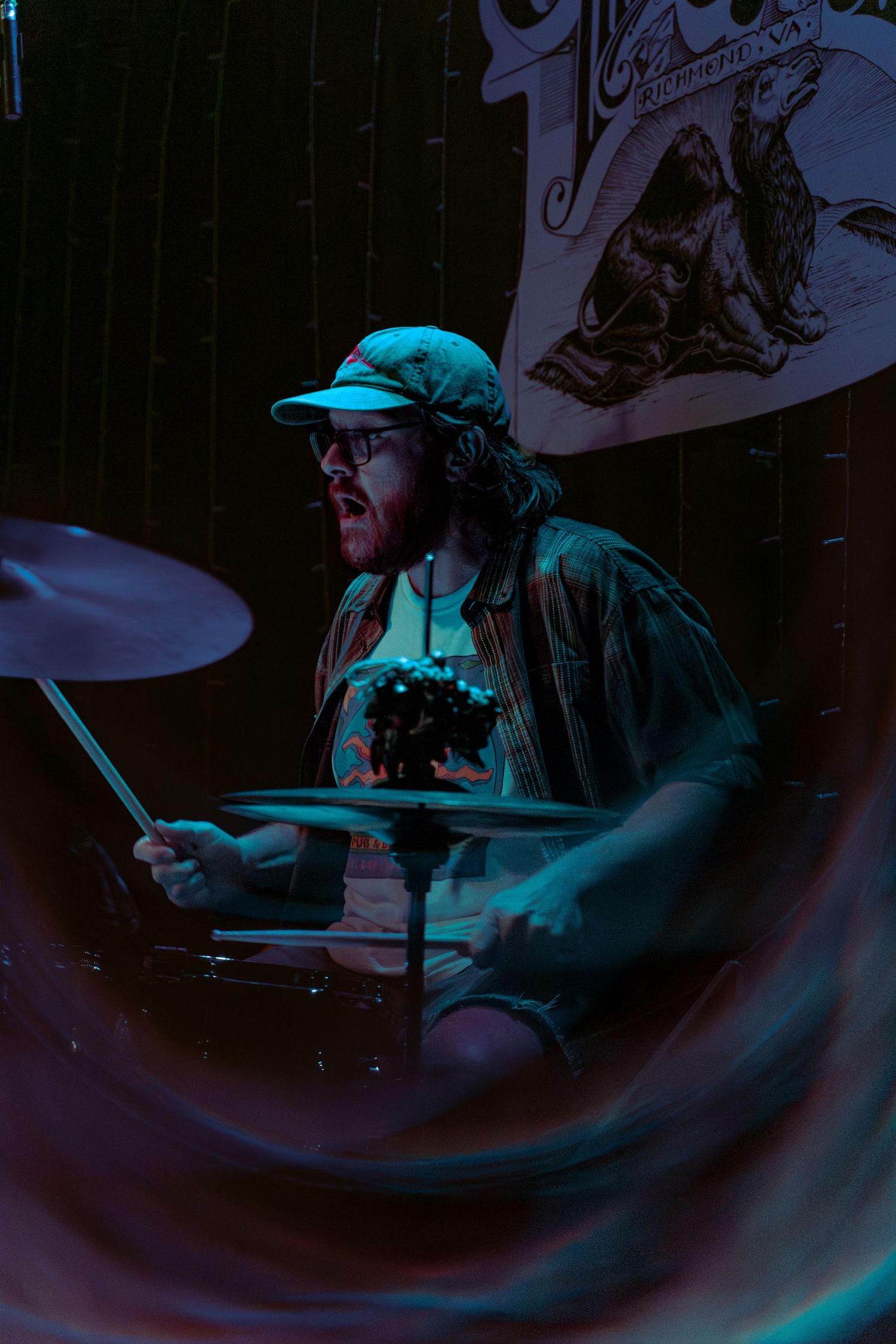 A drummer performs on stage lit by dramatic blue lighting while wearing a green baseball cap and glasses.