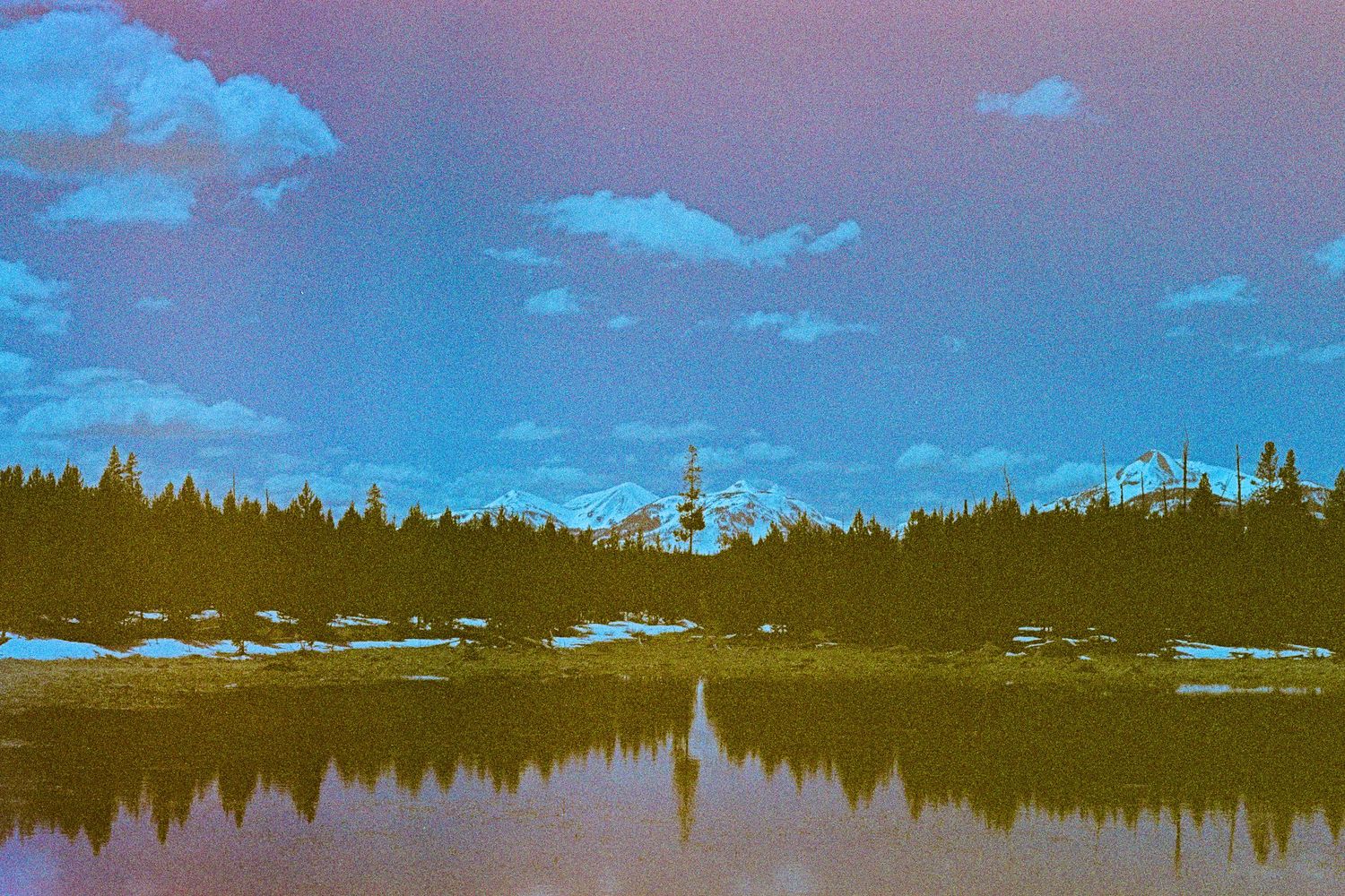 Snow-capped mountain peaks reflect in a calm lake surrounded by evergreen forest under a bright blue sky.