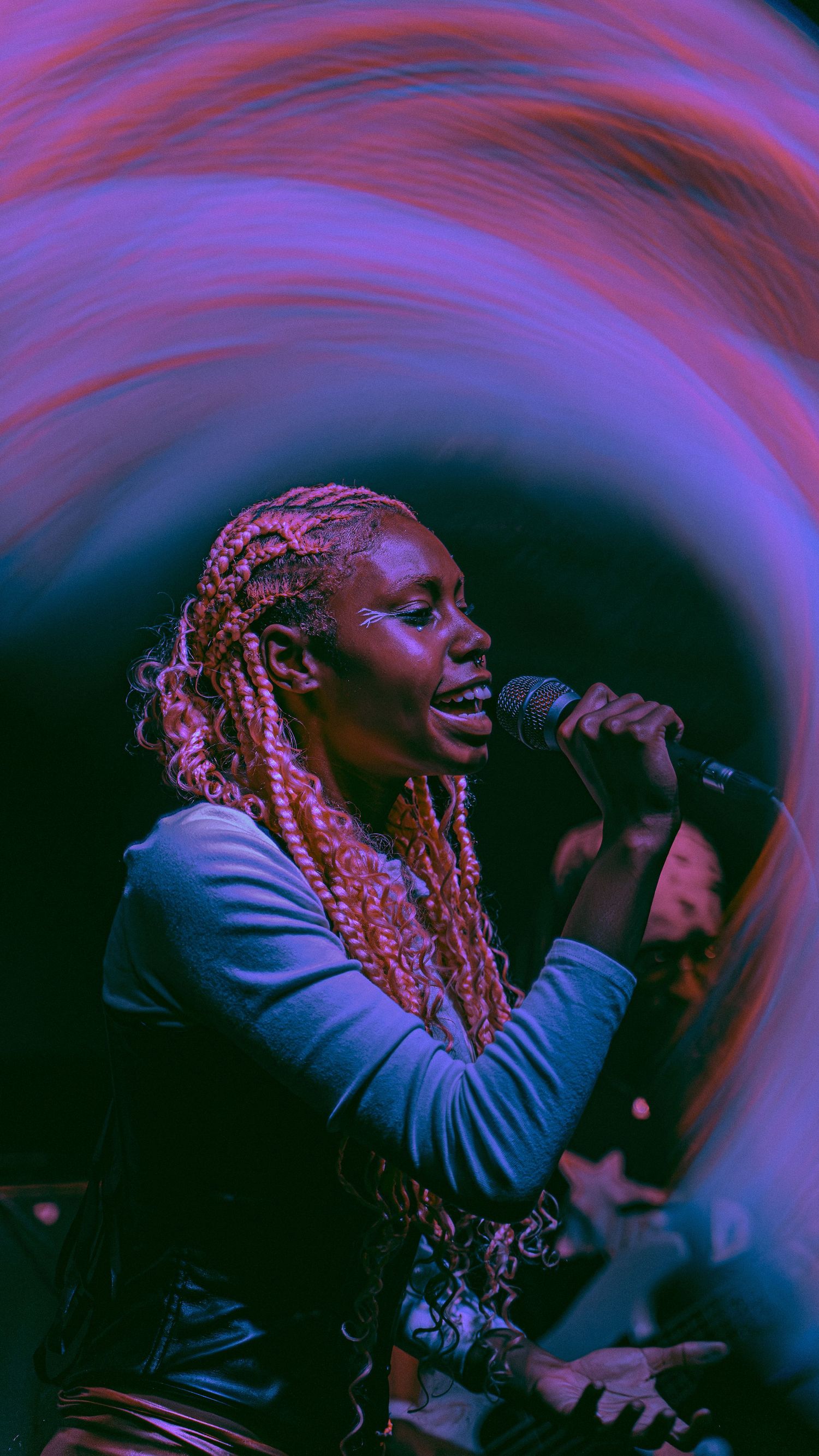 A performer sings into a microphone on stage with dramatic purple and pink lighting effects creating ethereal swirls above.