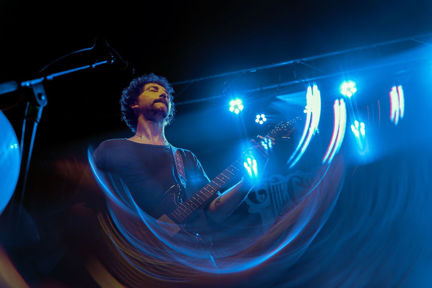 A performer on stage illuminated by blue lights creates ethereal light trails while singing into a microphone.