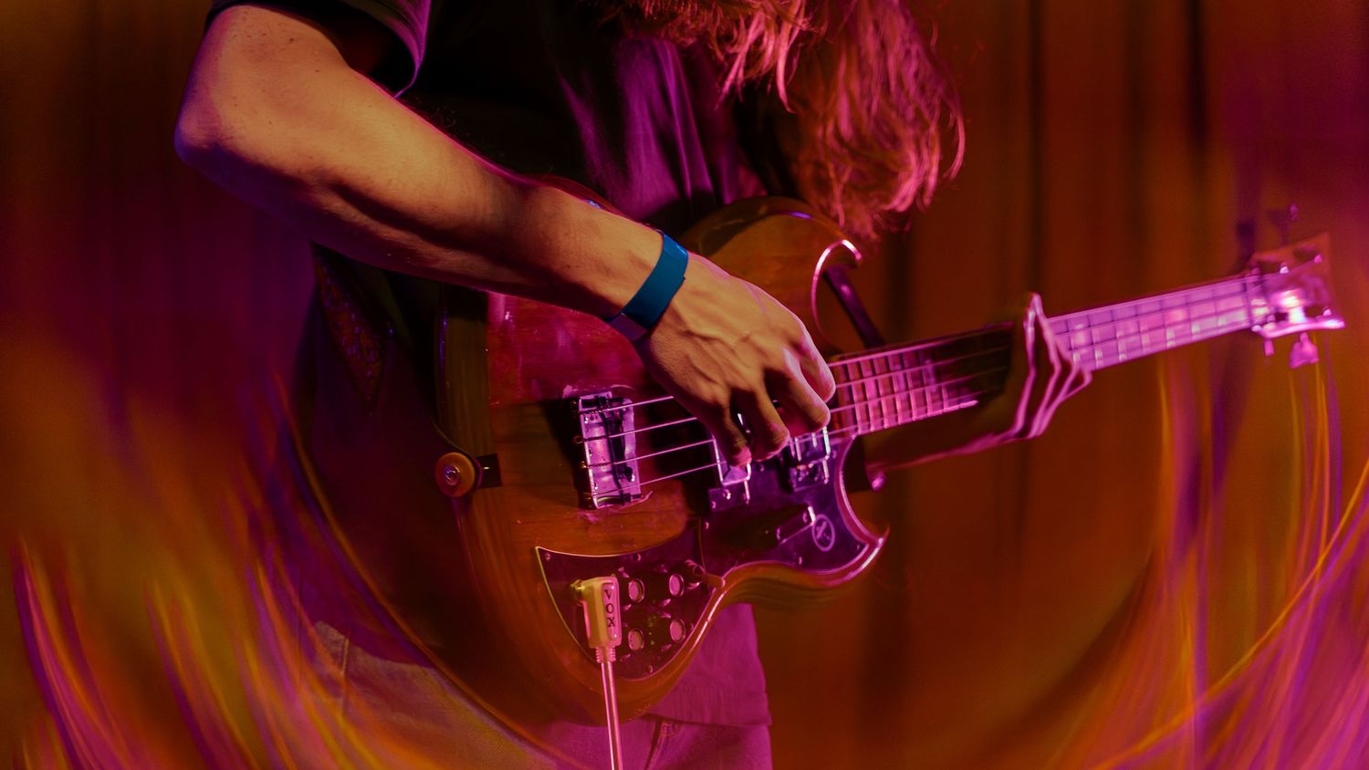 Musician plays a dark bass guitar on stage with vibrant red and orange lighting effects in the background.