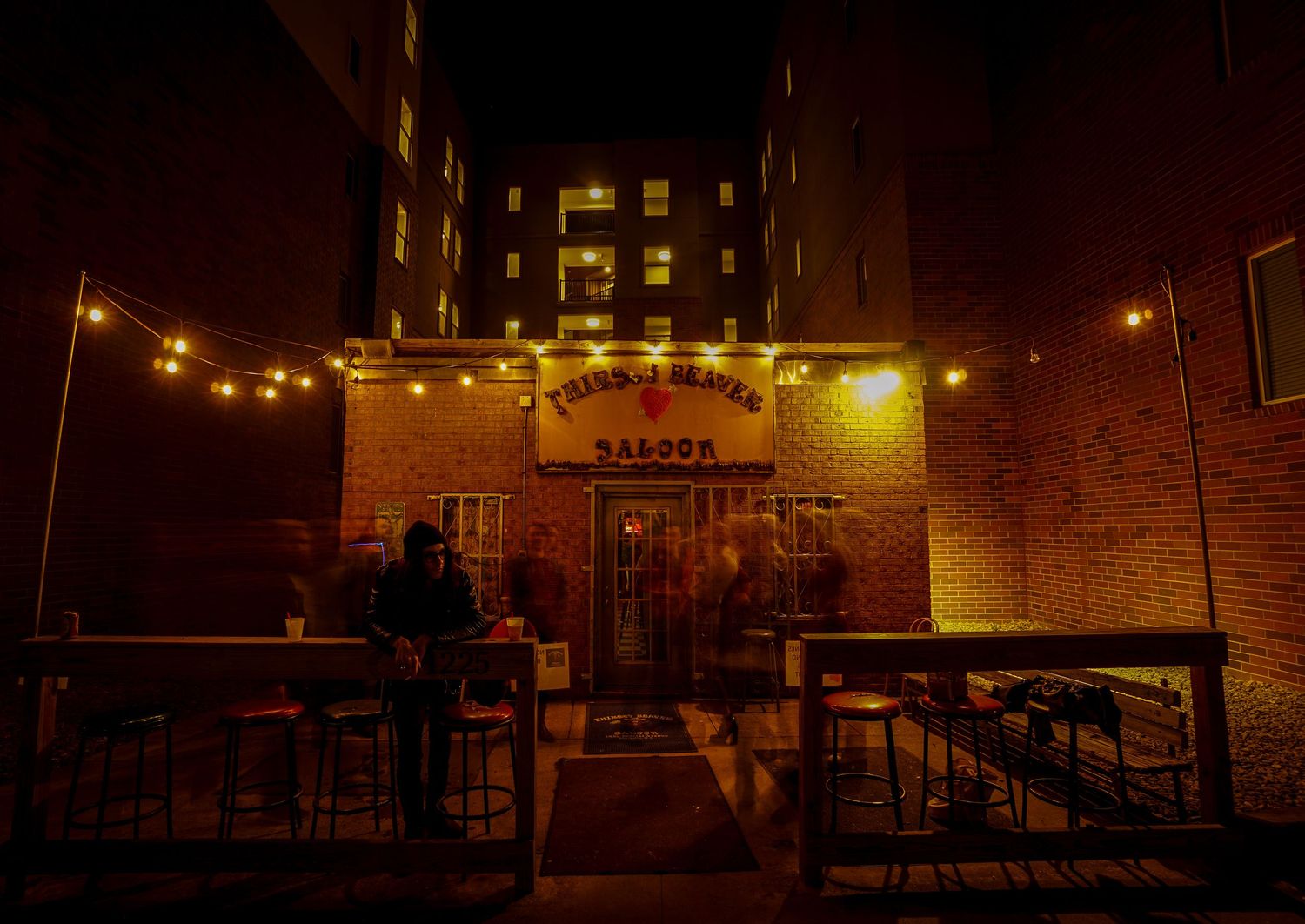 String lights illuminate a dark outdoor patio area with brick walls and wooden furniture.