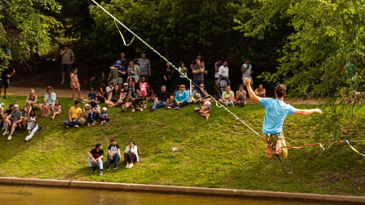 People watching a slackline performer cross over a grassy hill at an outdoor event.