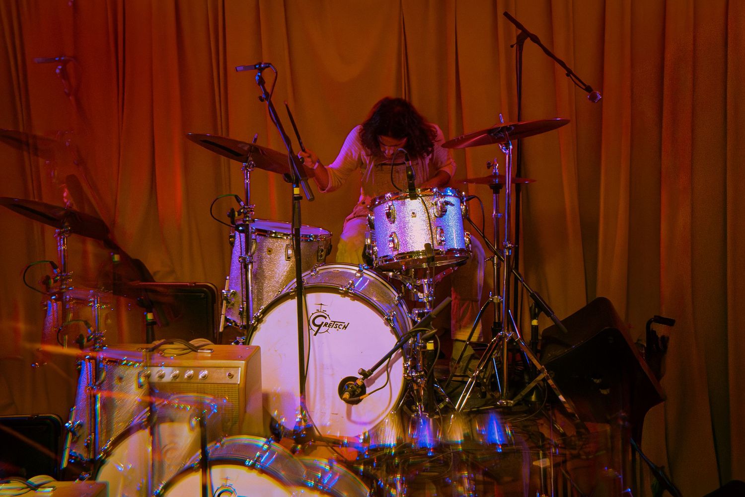 Musicians perform on stage with drums and instruments in a dimly lit venue with red and blue lighting.