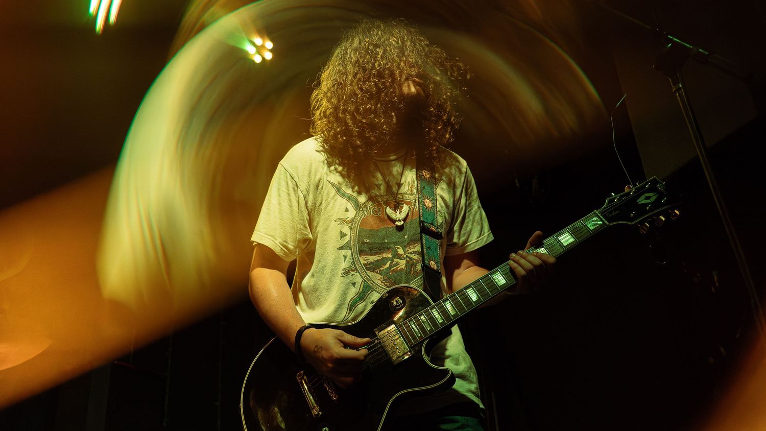 A guitarist performs on stage with curly hair and a white shirt, bathed in amber lighting while playing a performance.