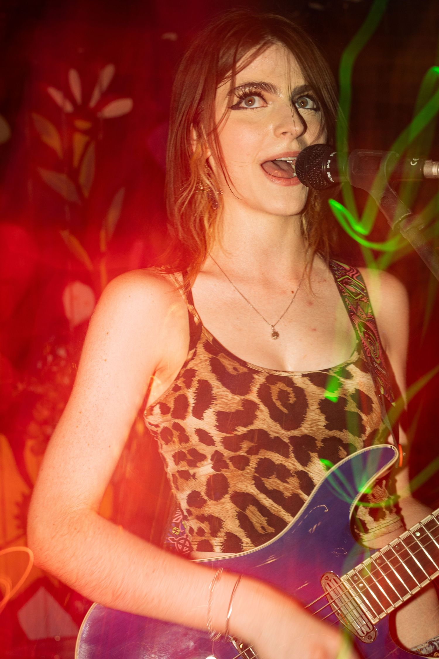 Performer in animal print tank top singing on stage with guitar under red and green lighting.