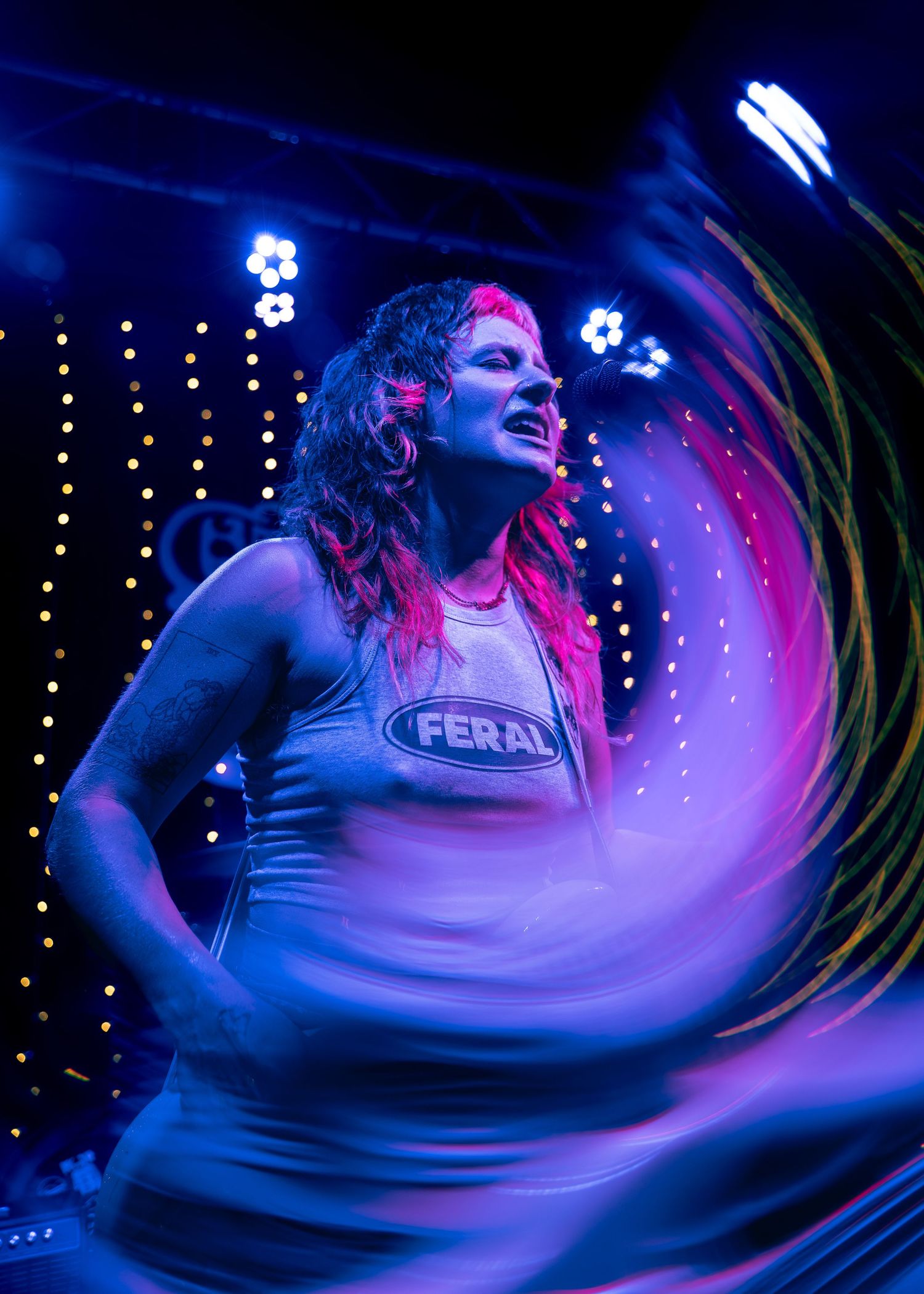 Musician wearing a Feral shirt performs on stage with vibrant red and blue lighting effects in the background.