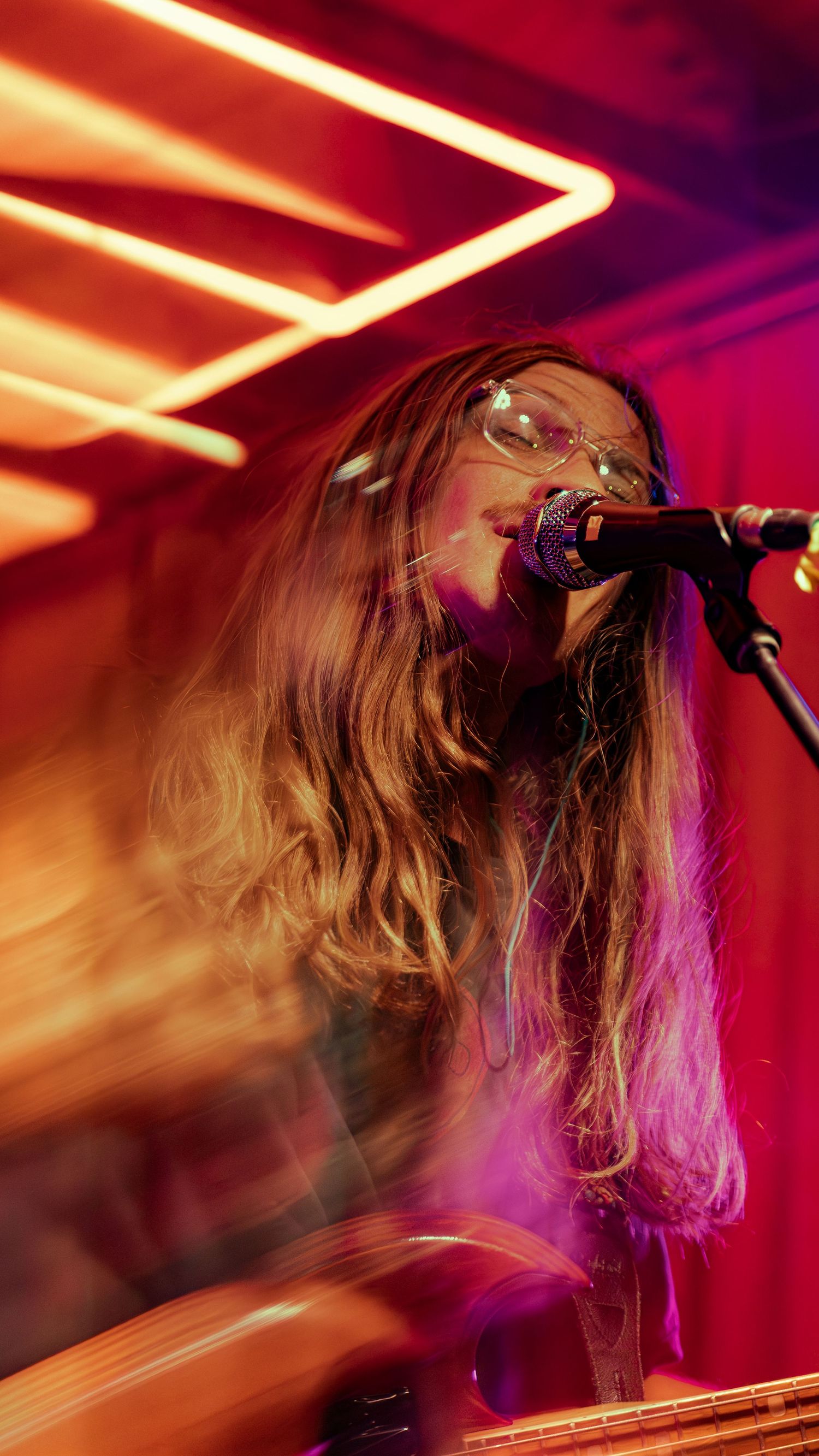 A dynamic concert performance captured in motion with vibrant red and purple stage lighting and dramatic hair movement.