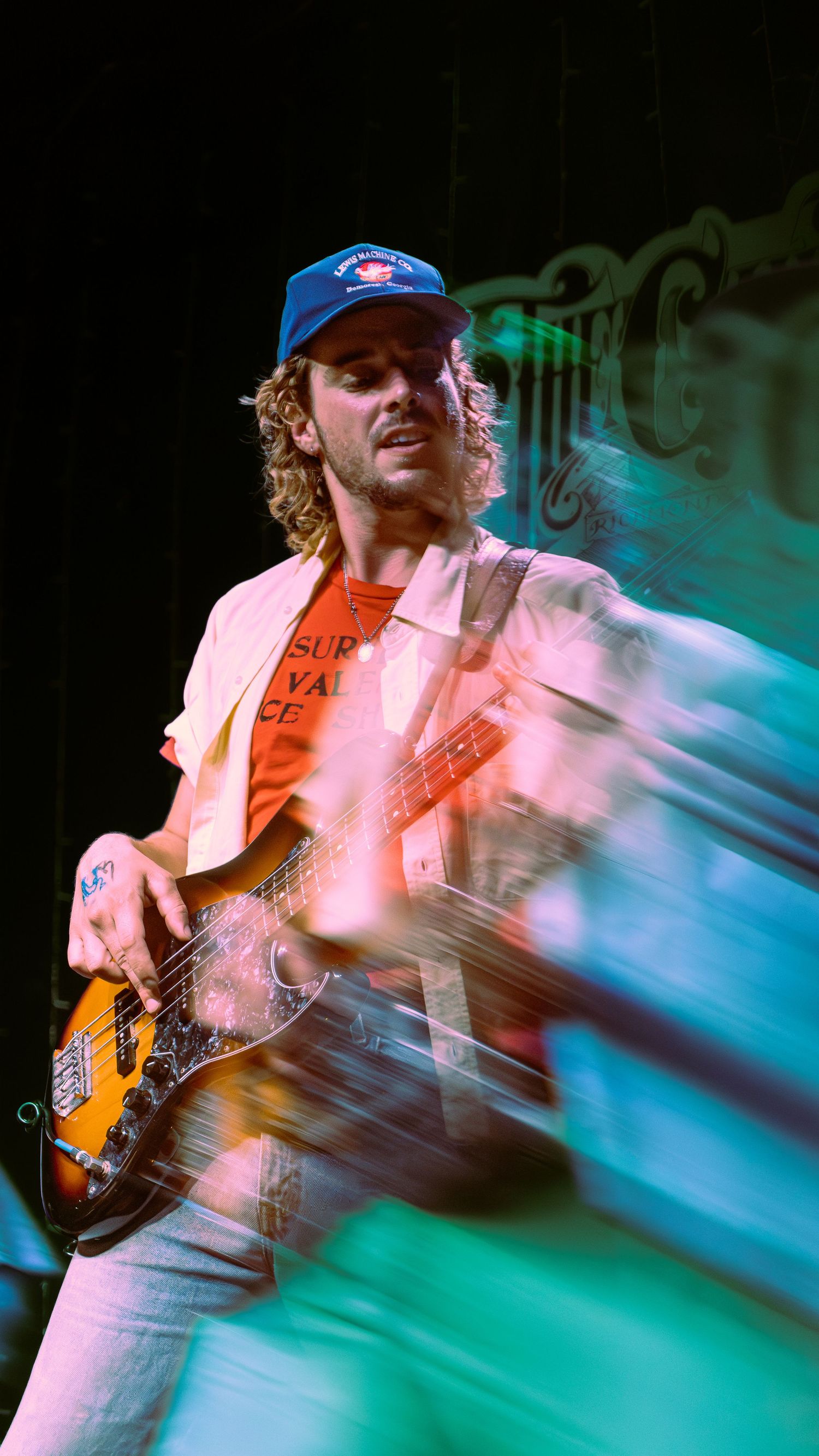 Musician wearing baseball cap plays bass guitar on stage with dramatic blue lighting and motion blur effects.