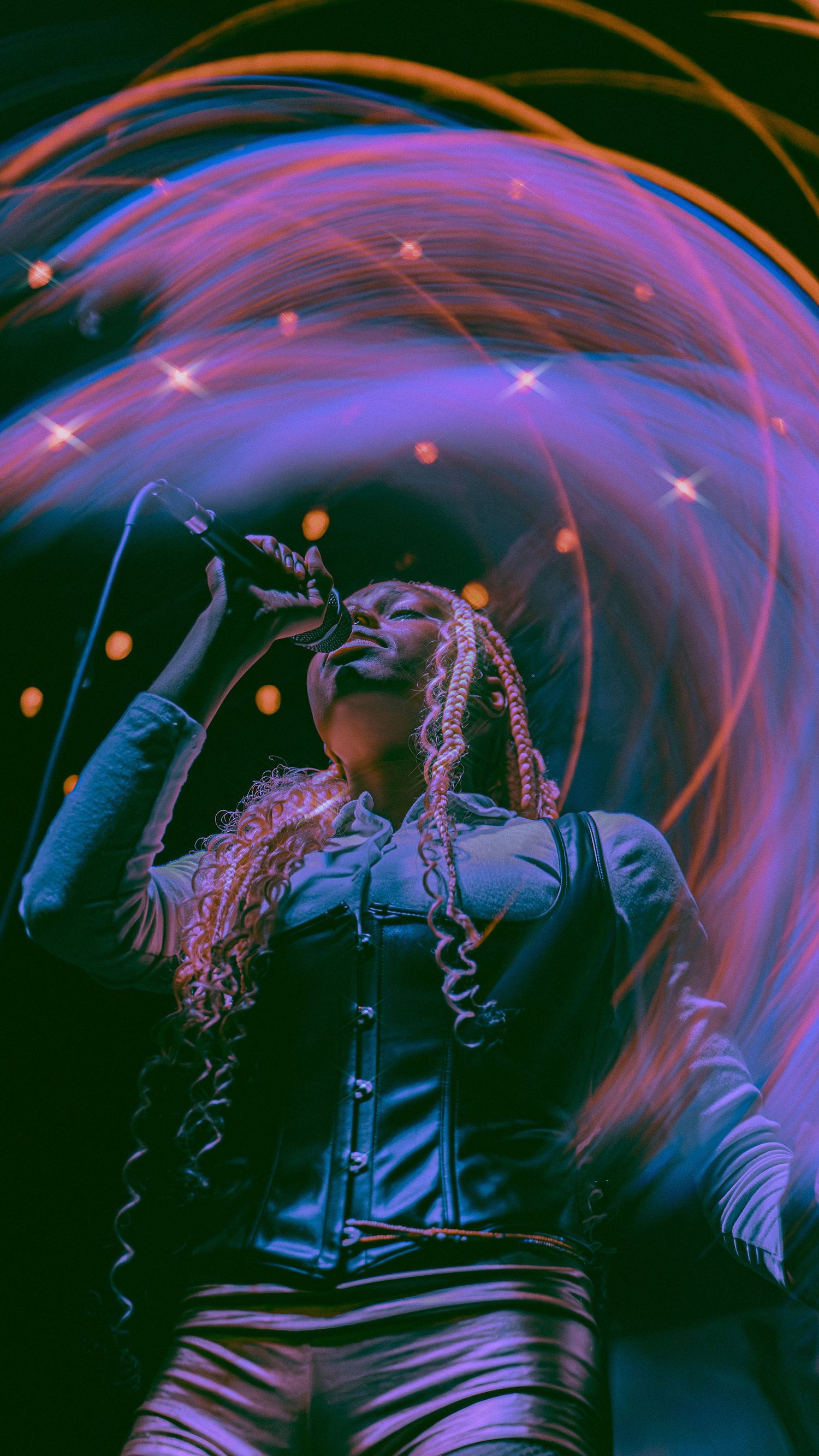 Concert performer on stage illuminated by swirling purple and orange lights creating dramatic light trails in a dark venue.