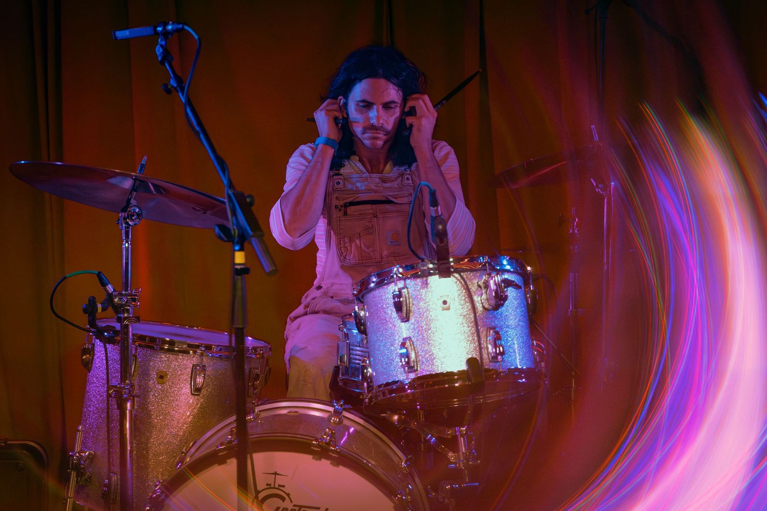 Drummer performs energetically behind a drum kit on stage with dramatic purple and red lighting effects.