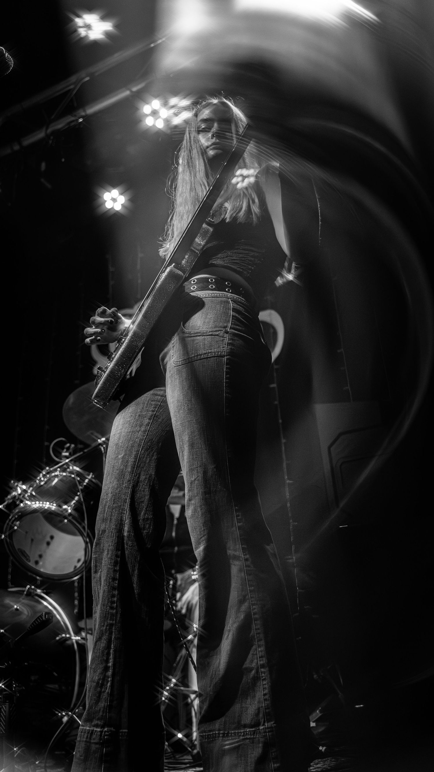 Black and white concert photo of a bassist performing on stage with flared jeans and dramatic lighting effects.