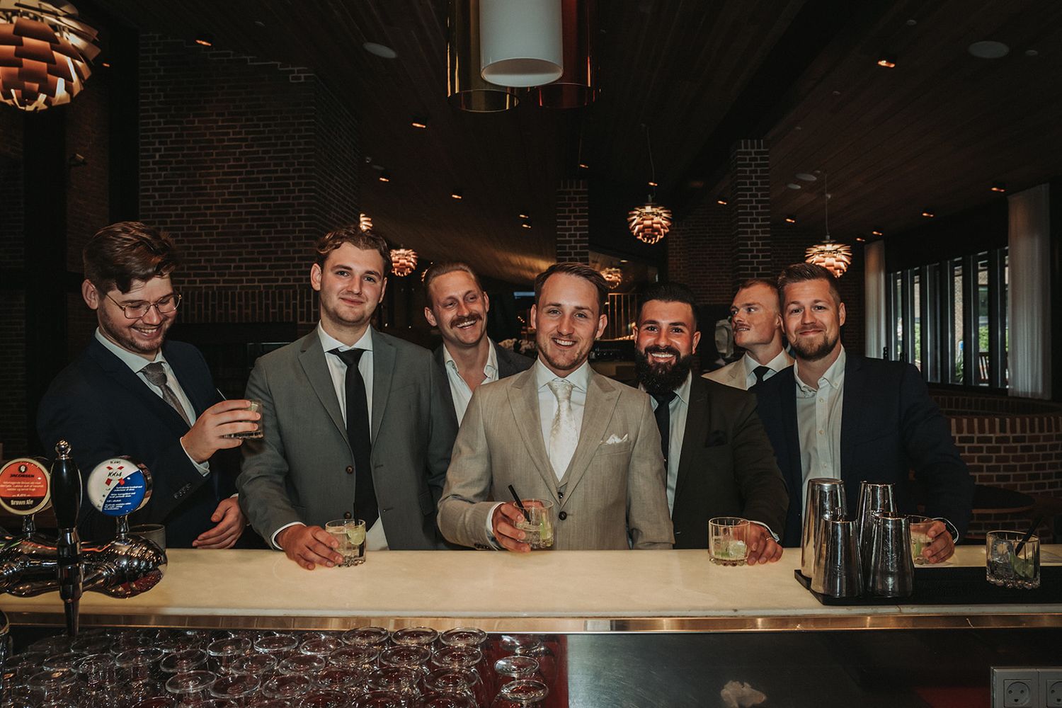 Groomsmen and wedding party members stand behind a wooden bar counter smiling and posing for a group photo.