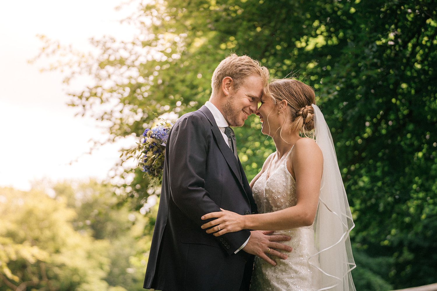 Couple sharing an intimate moment in a garden setting with sunlight filtering through trees.