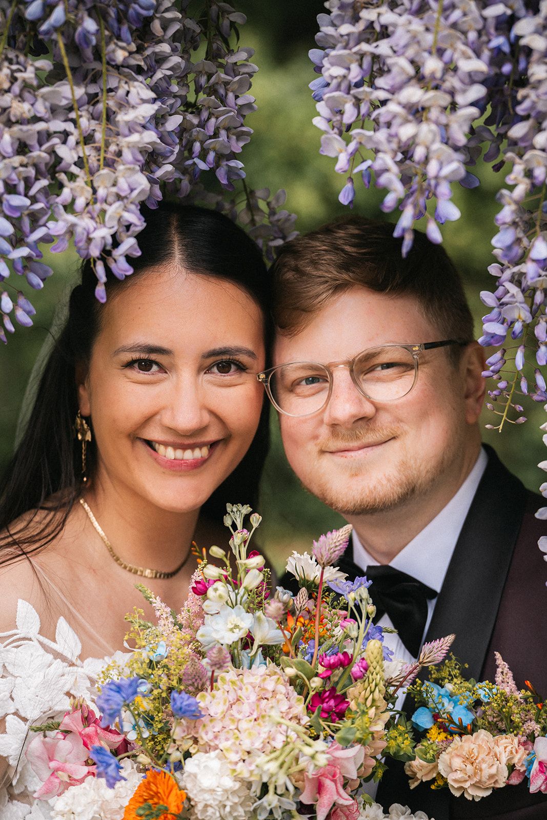 A couple posing under blooming wisteria with colorful wedding bouquet.