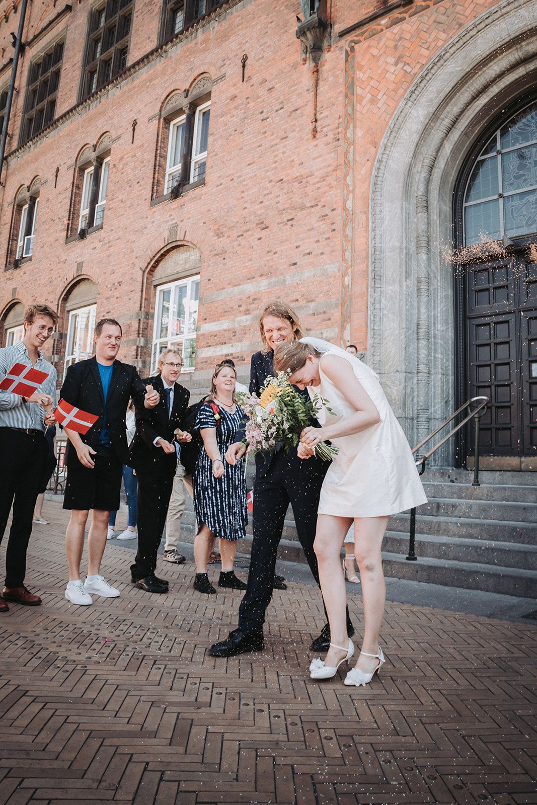 A small gathering celebrates outside a historic brick building with an arched doorway and Danish flag on a herringbone brick path.