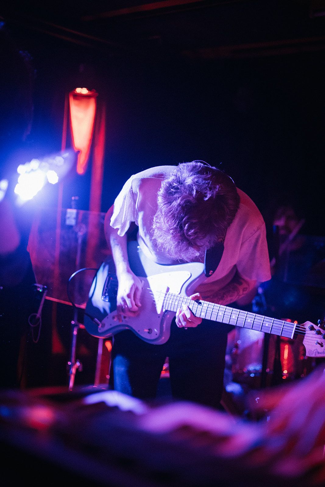 Musicians perform on stage in dramatic lighting with red and blue hues creating an atmospheric live music environment.