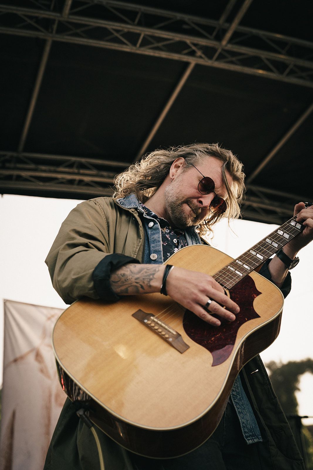 A musician with long hair and sunglasses performs with an acoustic guitar on an outdoor stage.