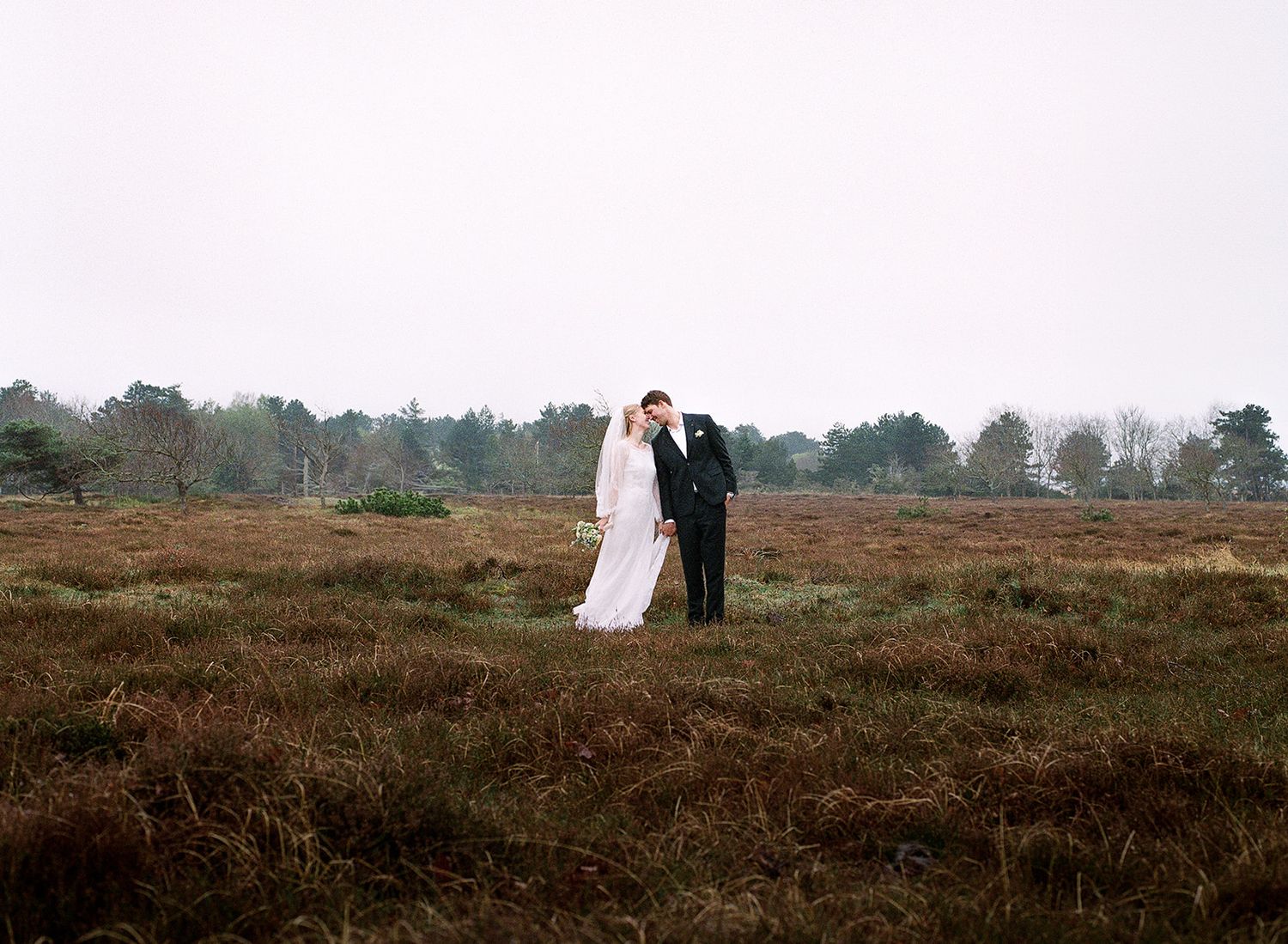 A couple in wedding attire stands together on a misty heathland with trees in the background.