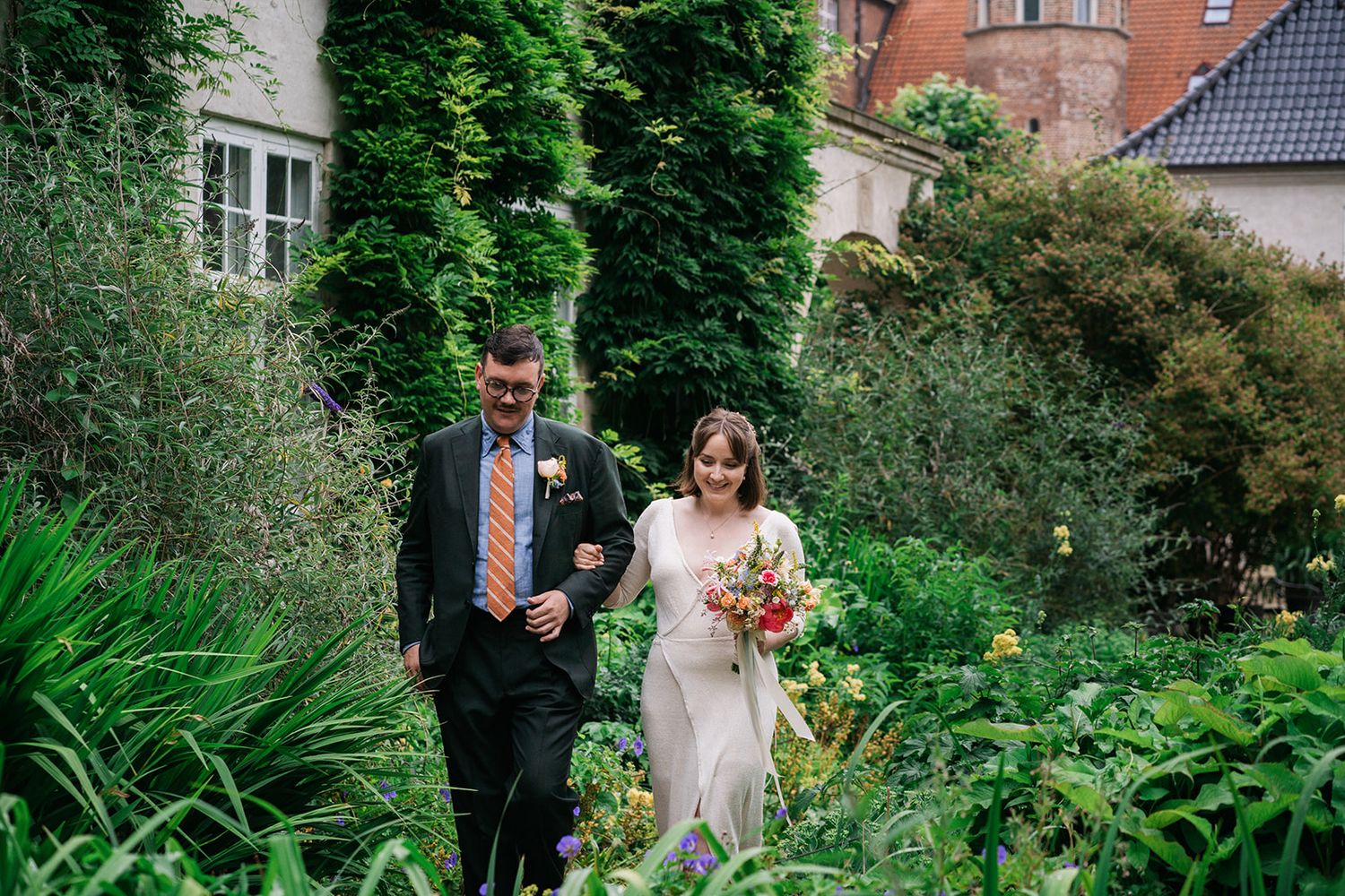 Two people walk along a garden path surrounded by lush greenery and flowers on a summer day.