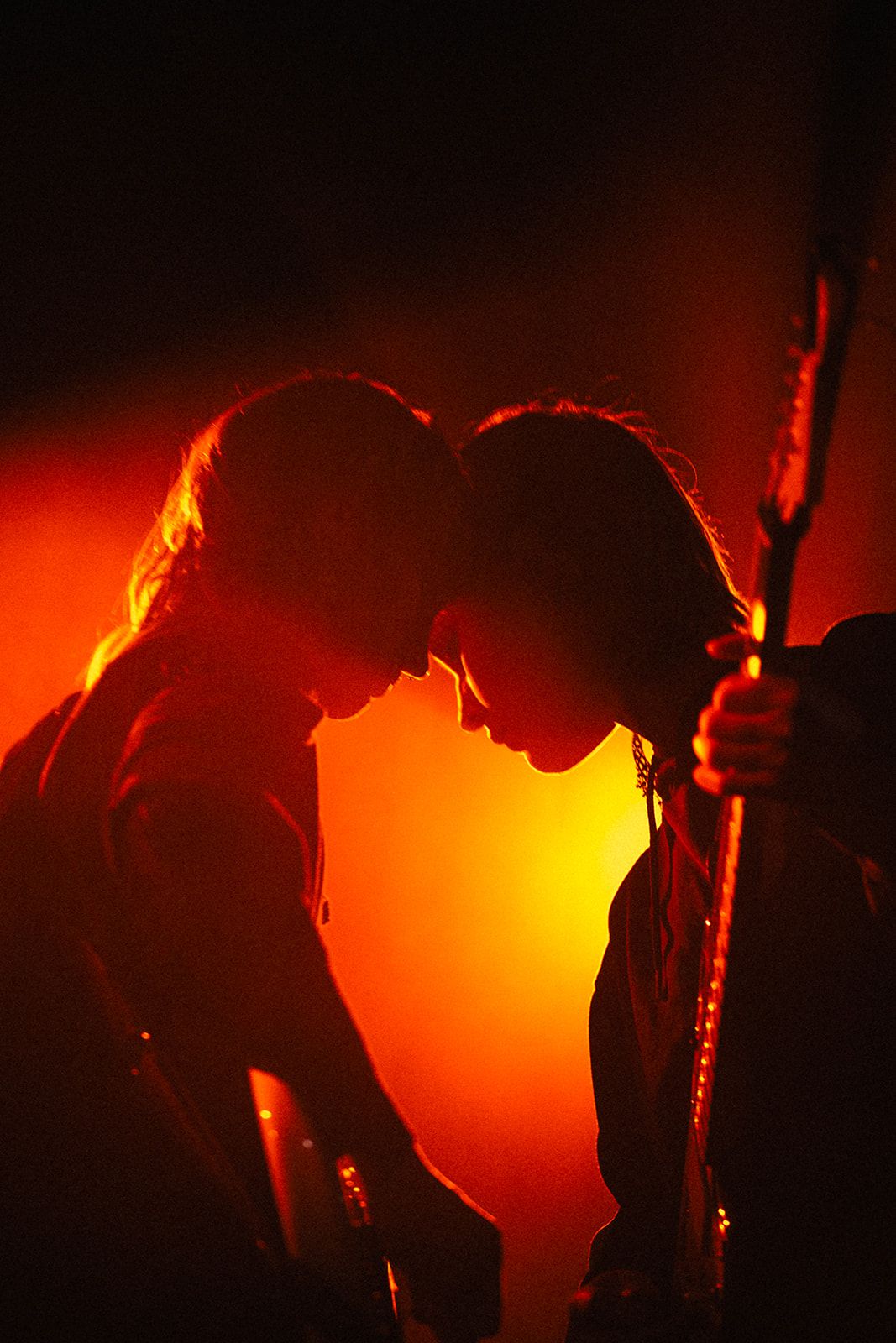 Silhouettes of musicians performing on stage are illuminated by dramatic orange and red backlighting creating a moody atmosphere.