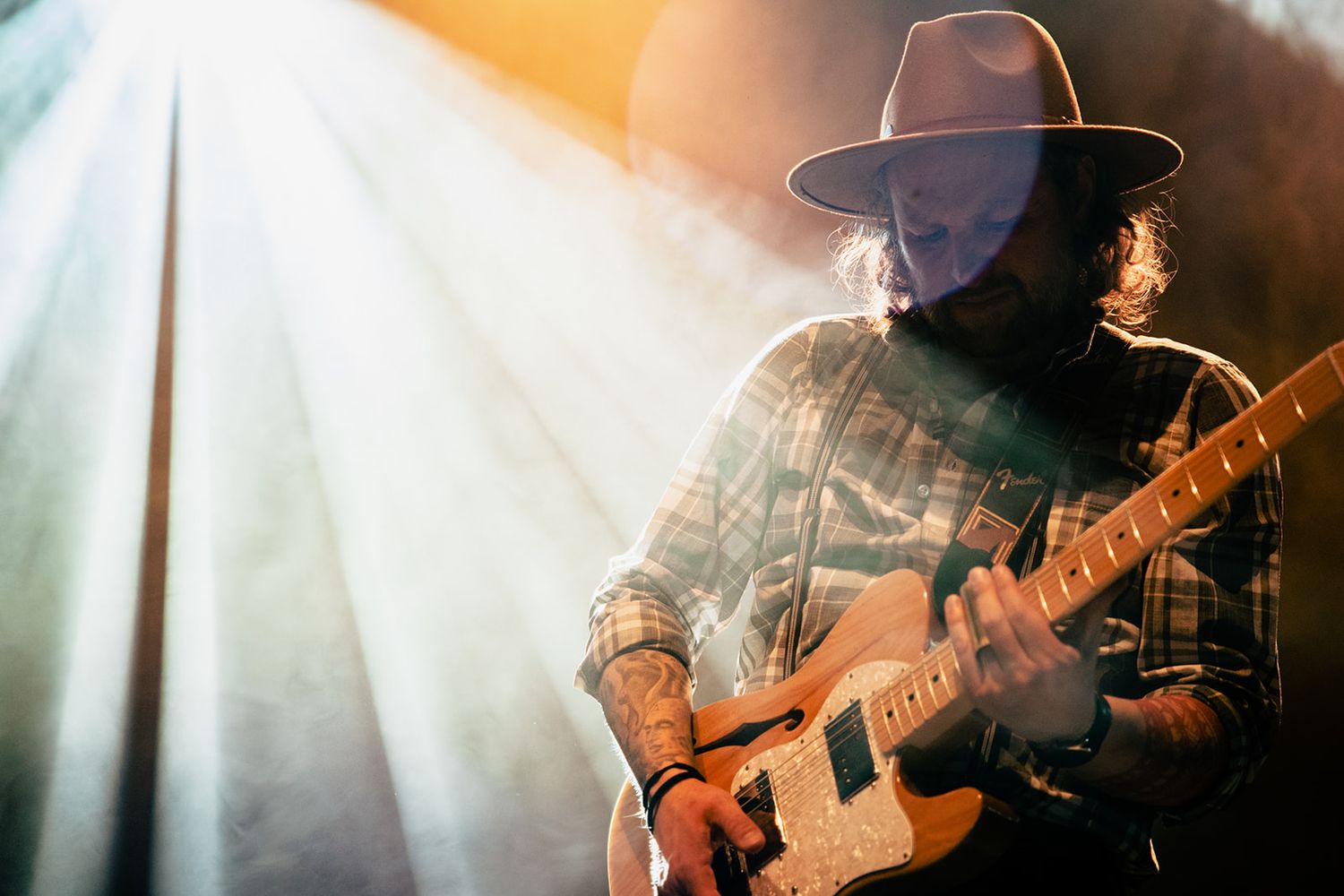 A guitarist in a western hat plays an electric guitar on stage with beams of light in the background.