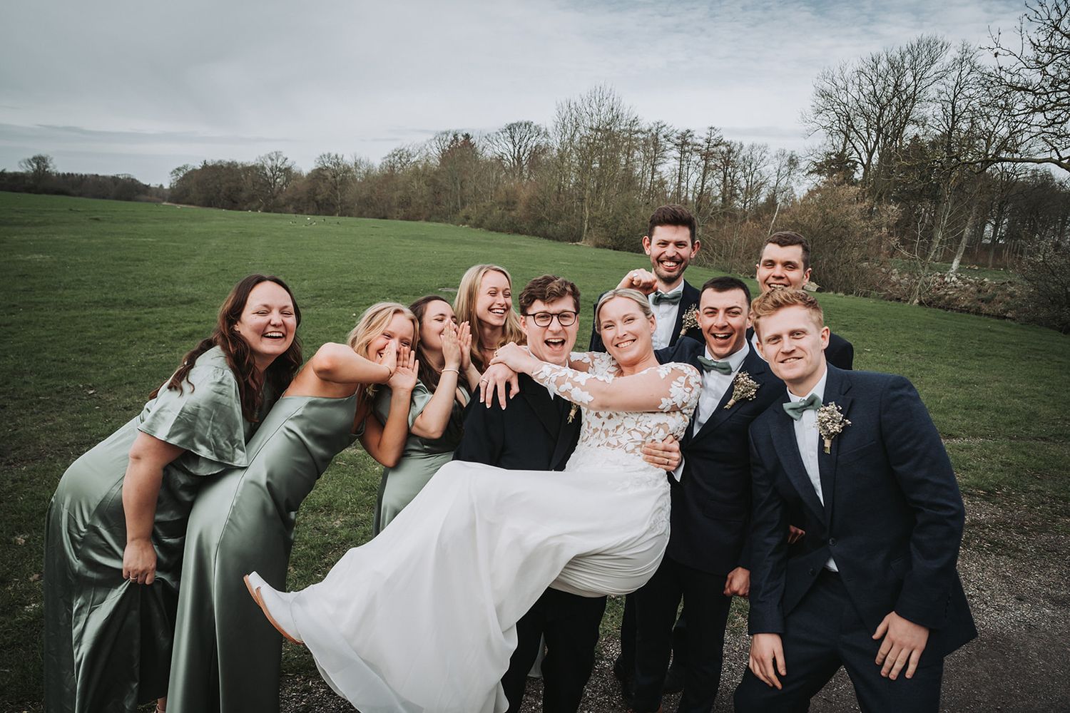 Wedding party poses and laughs together in a scenic grassy field during golden hour.