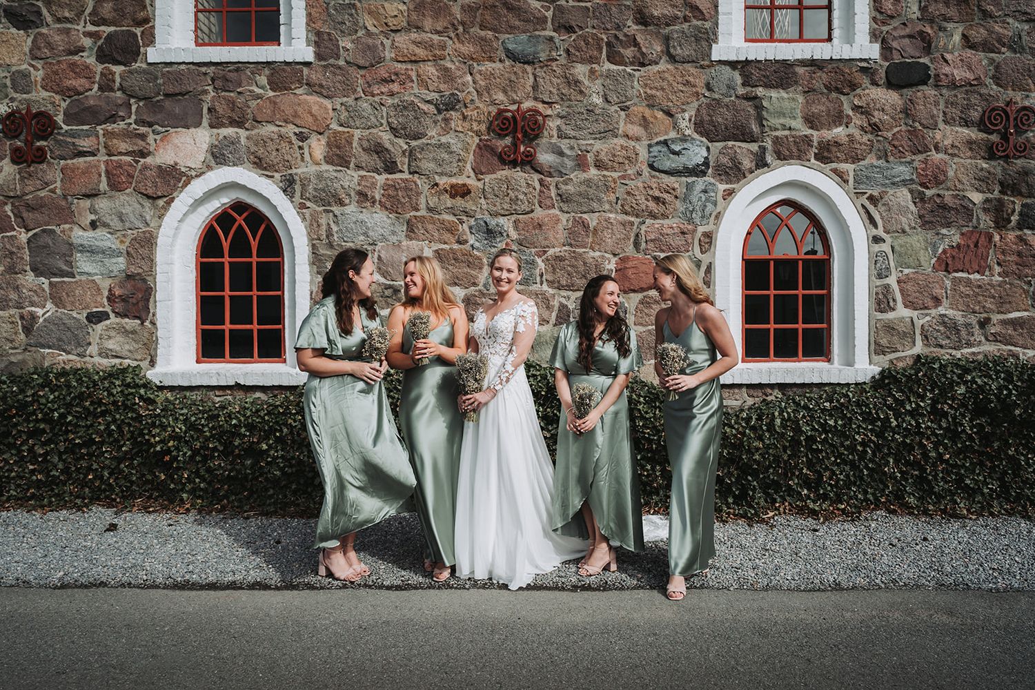 A wedding party sits together on stone steps outside a rustic building with large windows.