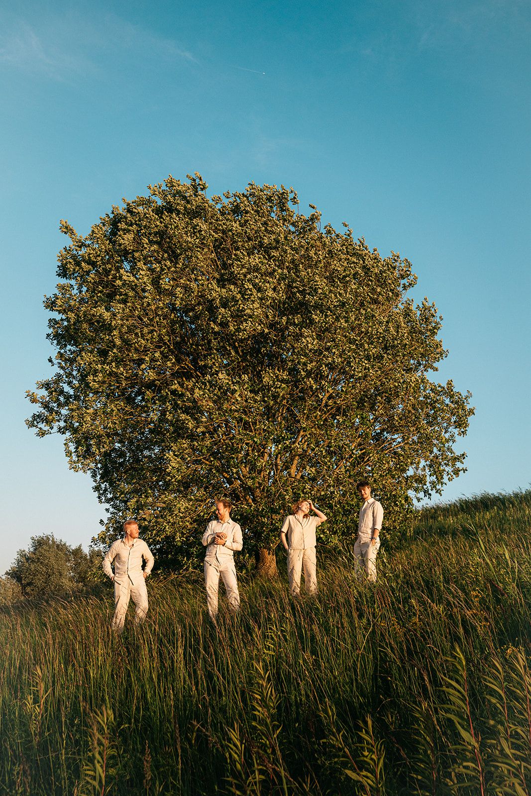 Four people in light colored outfits stand in a tall grass field at sunset with a large tree behind them.