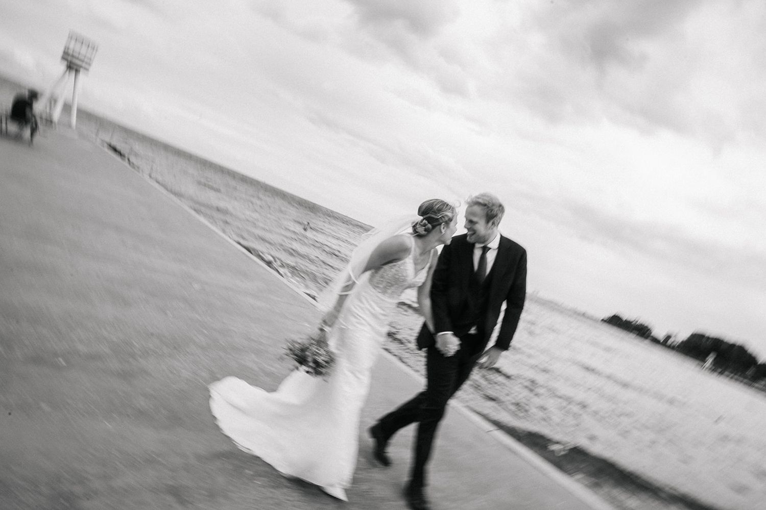 A romantic couple in formal wedding attire walks hand in hand along a scenic beach at sunset.